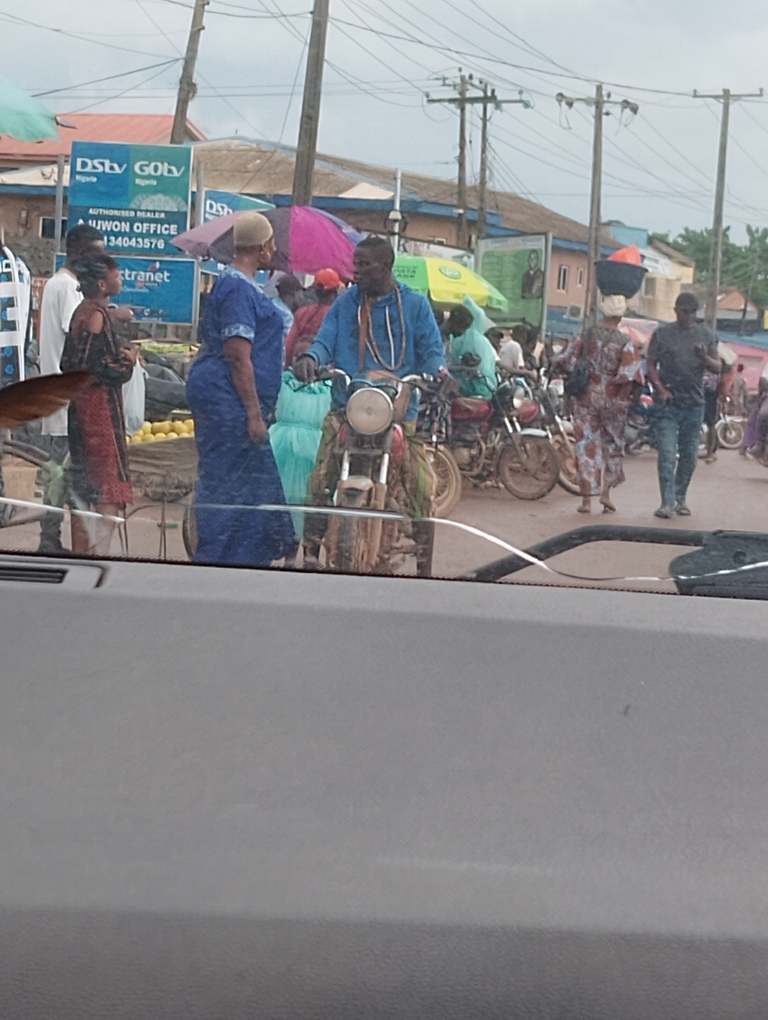 The image shows a busy street scene in Aketu-Oja, Nigeria. Several people are present, some standing and others on motorcycles. A man in a blue jacket is seated on a motorcycle, talking to a woman in a blue dress. Another woman, in a dark dress with red accents, stands nearby. In the background, a sign advertising DStv and GOtv is visible. Several motorcycles are parked or moving on the street. The environment appears to be an outdoor market or roadside area with various stalls and shops. Overhead power lines and utility poles are also present. The ground is a dirt or paved road. The sky is overcast.