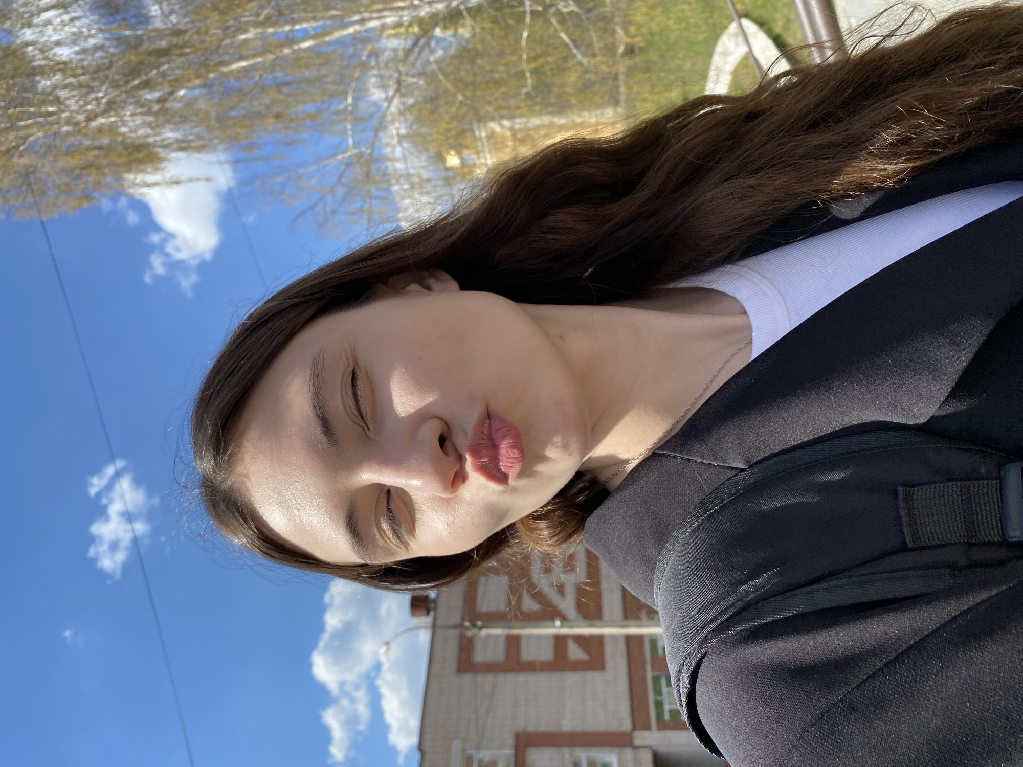 A young woman with long brown hair is posing for a selfie outdoors. She has fair skin, closed eyes, and is puckering her lips. She is wearing a white top and a black jacket with a dark-colored backpack strap visible over her shoulder. The background consists of a clear blue sky with scattered white clouds, and the tops of trees with bare branches can be seen. A building with a light-colored facade and red brick accents is partially visible in the lower portion of the frame. The photograph appears to be taken in Votkinsk, Russia, as identified by the building's architecture and the presence of Cyrillic signage.