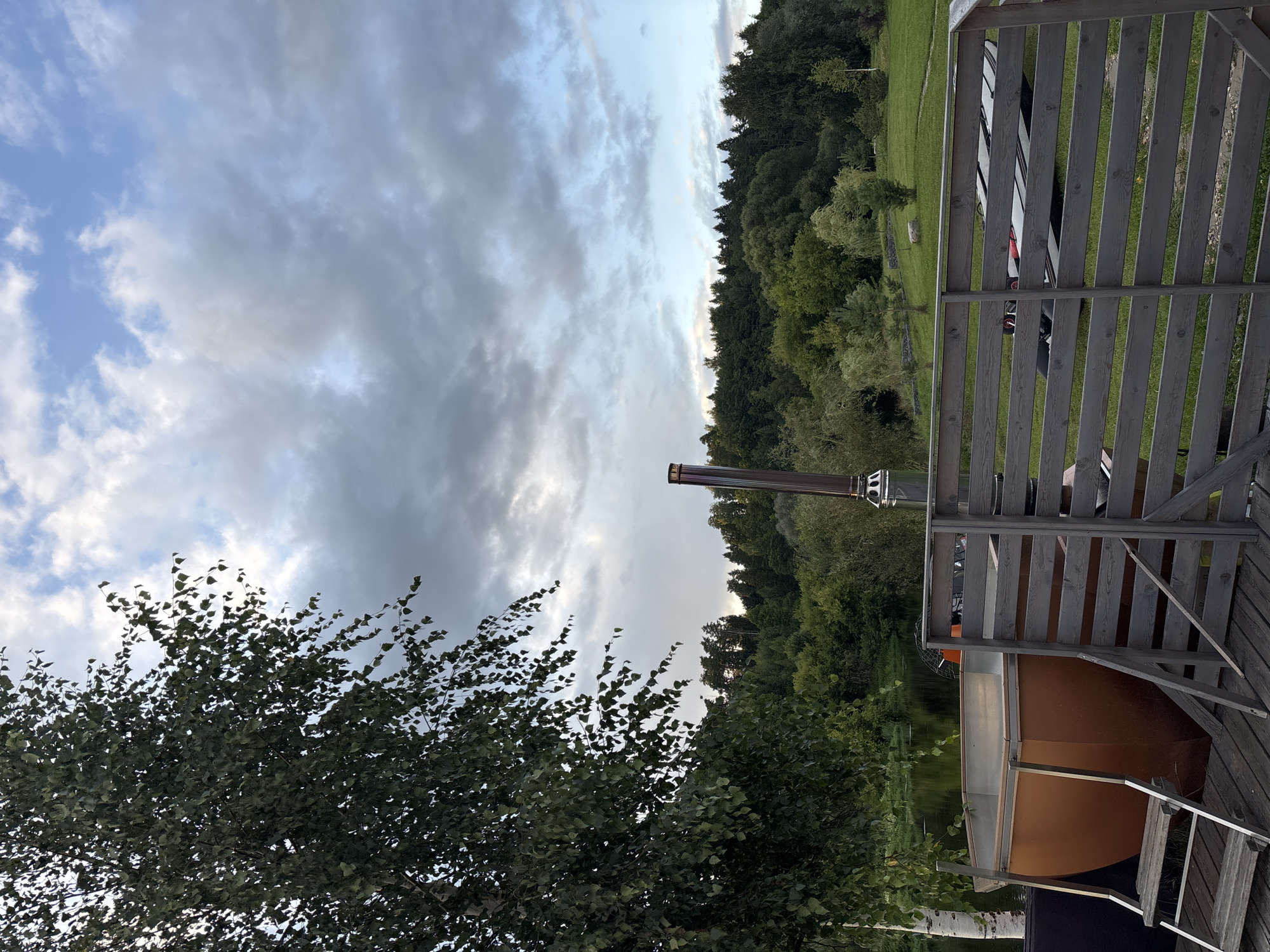 The image depicts an outdoor scene with a hot tub on a wooden deck in Zvenigorod, Russia. A tall birch tree with dark green foliage dominates the left side of the frame, reaching towards a cloudy sky with patches of blue. To the right of the hot tub, a wooden fence encloses part of the deck, where a long, white object with red accents, possibly a surfboard or a paddleboard, is visible. Beyond the deck, a calm body of water, likely a river, is visible, bordered by a dense forest of green trees. The sky is overcast with heavy, grey clouds interspersed with lighter, brighter areas, suggesting either early morning or late afternoon lighting. The overall atmosphere is serene and natural.