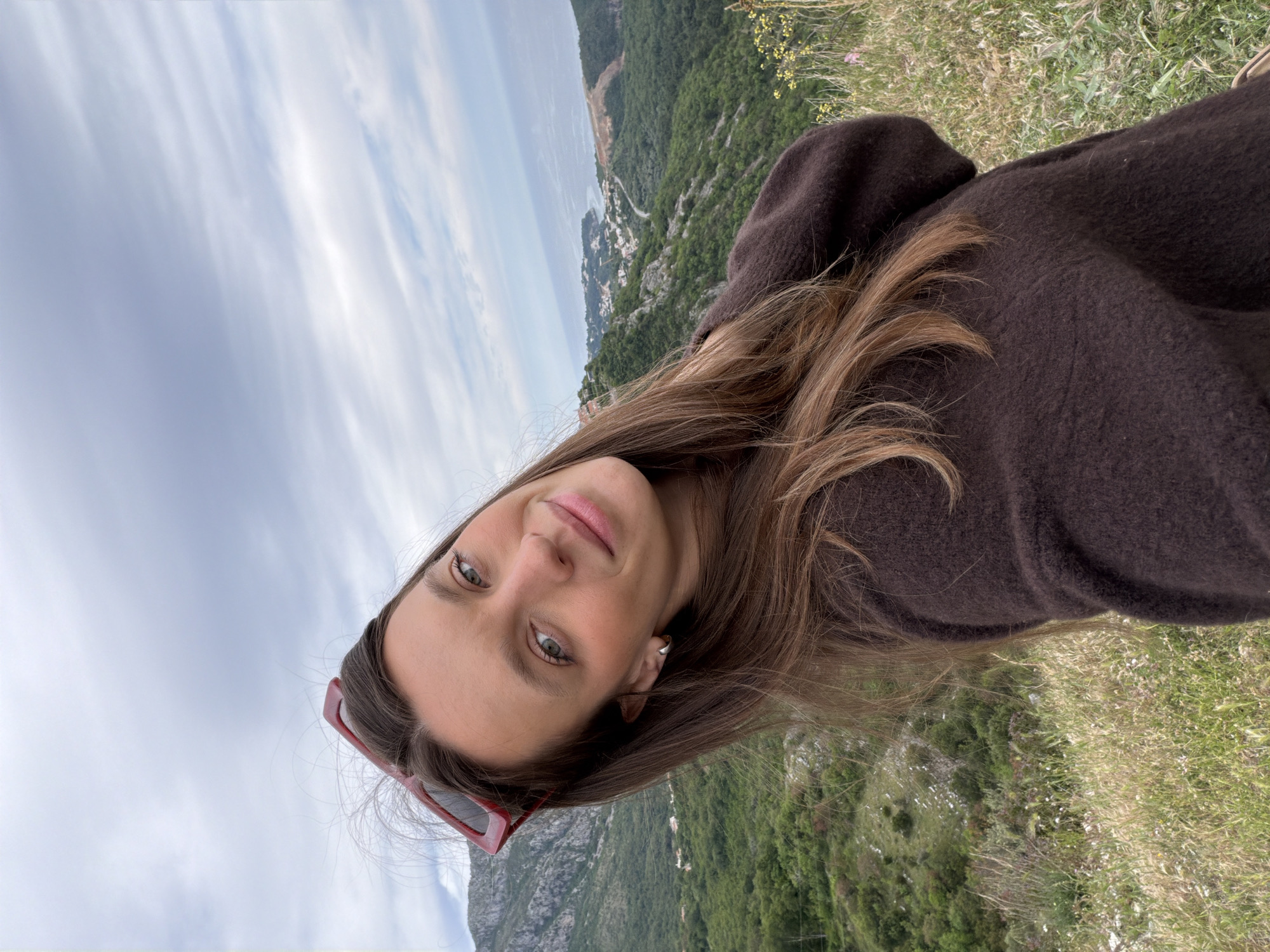 A woman is taking a selfie outdoors. She has long brown hair and is wearing a brown hooded sweatshirt. Red sunglasses are perched on her head. In the background is a mountainous landscape with lush green vegetation and a town nestled near the coast. The sky is overcast. The location appears to be near Sveti Stefan, Montenegro, as inferred by the coastal scenery and mountainous terrain.