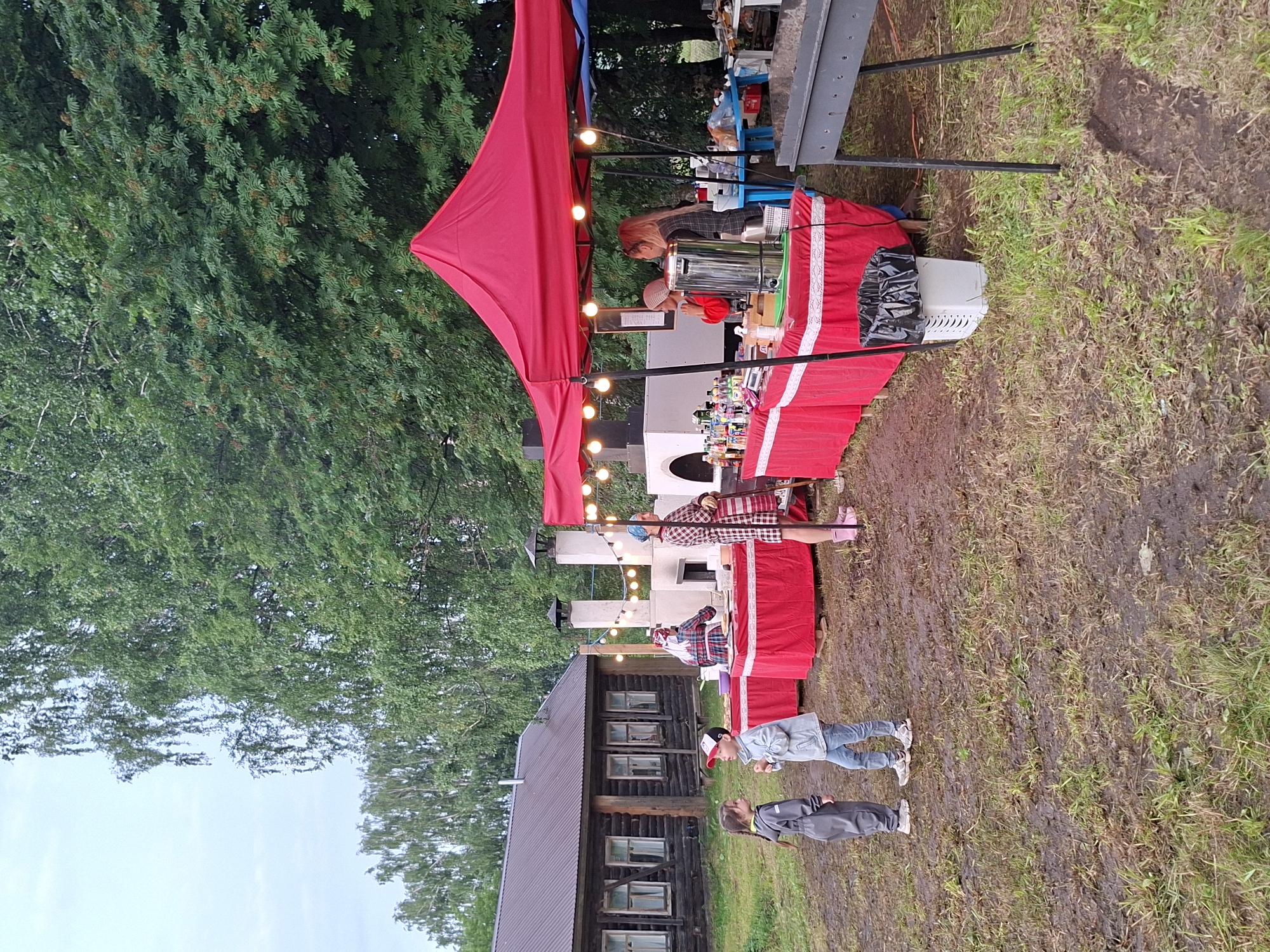 The image depicts an outdoor market or festival scene set in Старые Быги, Russia. A large red canopy tent is set up, adorned with string lights, suggesting an evening or overcast day. Several people are visible, some interacting with a stall that appears to be selling drinks or food, with bottles and dispensers displayed. A woman in a plaid shirt is working behind the counter, and another person in a similar outfit is also near the stall. Further down, two children are standing near a rustic wooden building. The ground is muddy, indicating recent rain or damp conditions, and surrounded by lush green trees. The overall atmosphere seems relaxed and community-oriented. No visible text is present in the image.