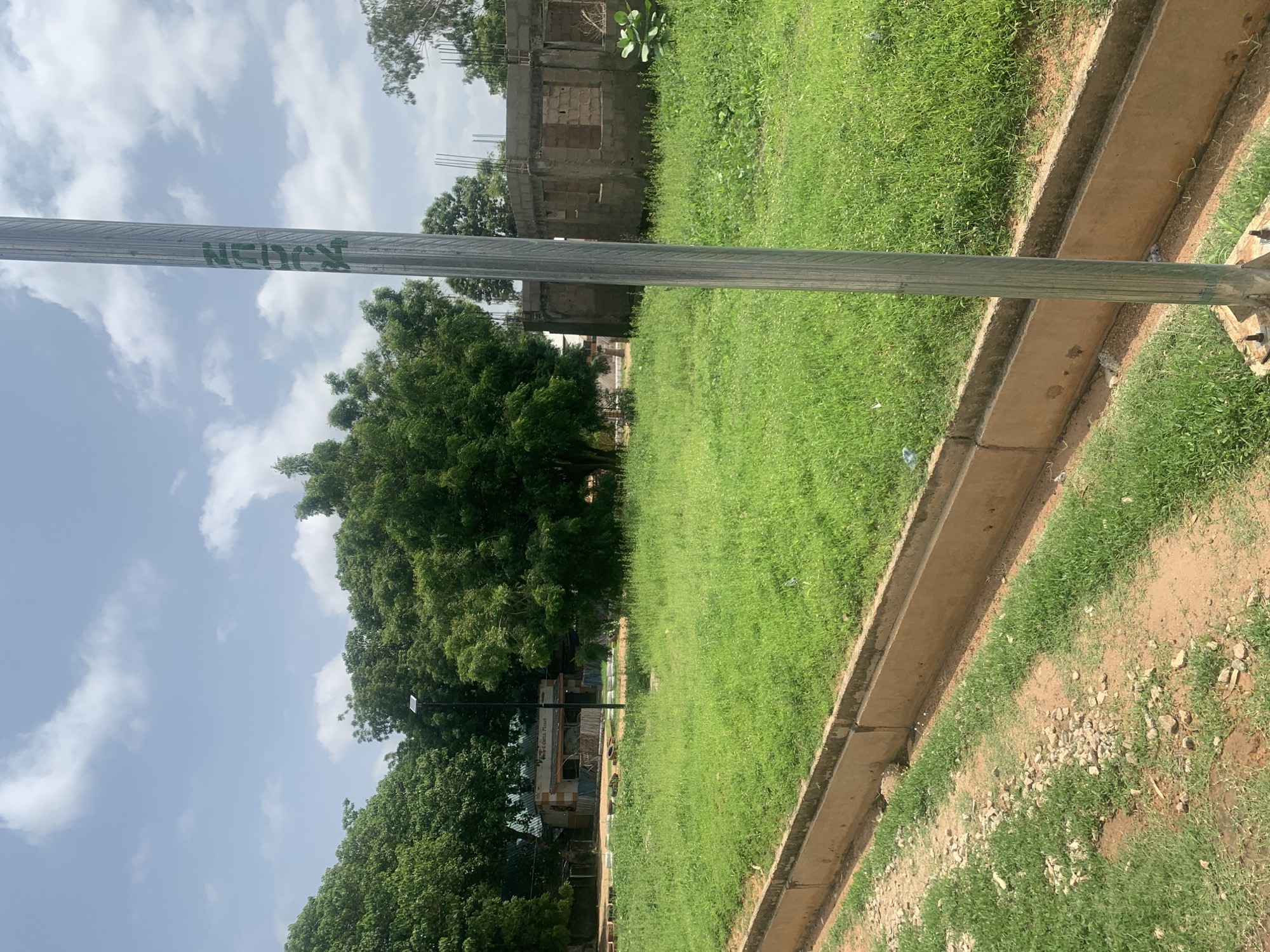 The image depicts an outdoor scene on a sunny day in Jalingo, Nigeria, possibly near a road. In the foreground, a gravelly dirt path runs alongside a concrete drainage channel filled with green grass. A tall, corrugated metal pole stands vertically in the center of the frame, marked with green lettering that reads "MON 14". Beyond the grassy median strip, a cluster of lush green trees dominates the middle ground. To the left, behind the trees, a low-rise building with a light-colored facade and a dark roof is visible, suggesting a commercial establishment. To the right, a partially constructed building made of concrete blocks stands open with visible rebar, indicating ongoing construction. The sky above is partly cloudy with fluffy white clouds against a light blue backdrop.