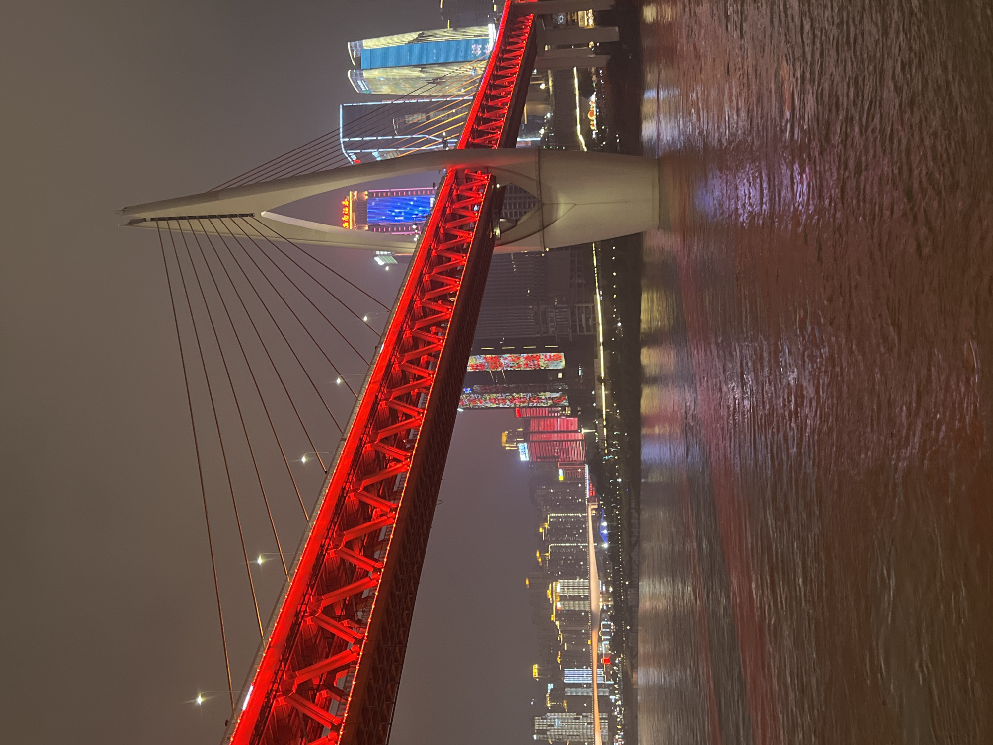 An eye-level, vertical, medium shot captures the Hongyadong Bridge at night in Yuzhong District, China. The bridge, illuminated with vibrant red lights, stretches across the frame from left to right, its steel lattice structure visible. A massive, white cable-stayed tower, a defining feature of the bridge, rises on the right side of the image, supporting the red deck with taut cables. The dark, cloudy sky above is dotted with streetlights and the warm glow of the cityscape.
In the background, the Yuzhong District skyline is a tapestry of illuminated skyscrapers, displaying a variety of colors, with a prominent blue-lit building standing out. The Jialing River flows in the foreground, its surface reflecting the colorful lights of the bridge and the city, creating a reddish-brown hue with ripples and waves. The water's surface also shows a distinct red stripe, likely a reflection of the bridge's illumination. No people are visible in the scene. The image is taken at night, with artificial lights dominating the scene.