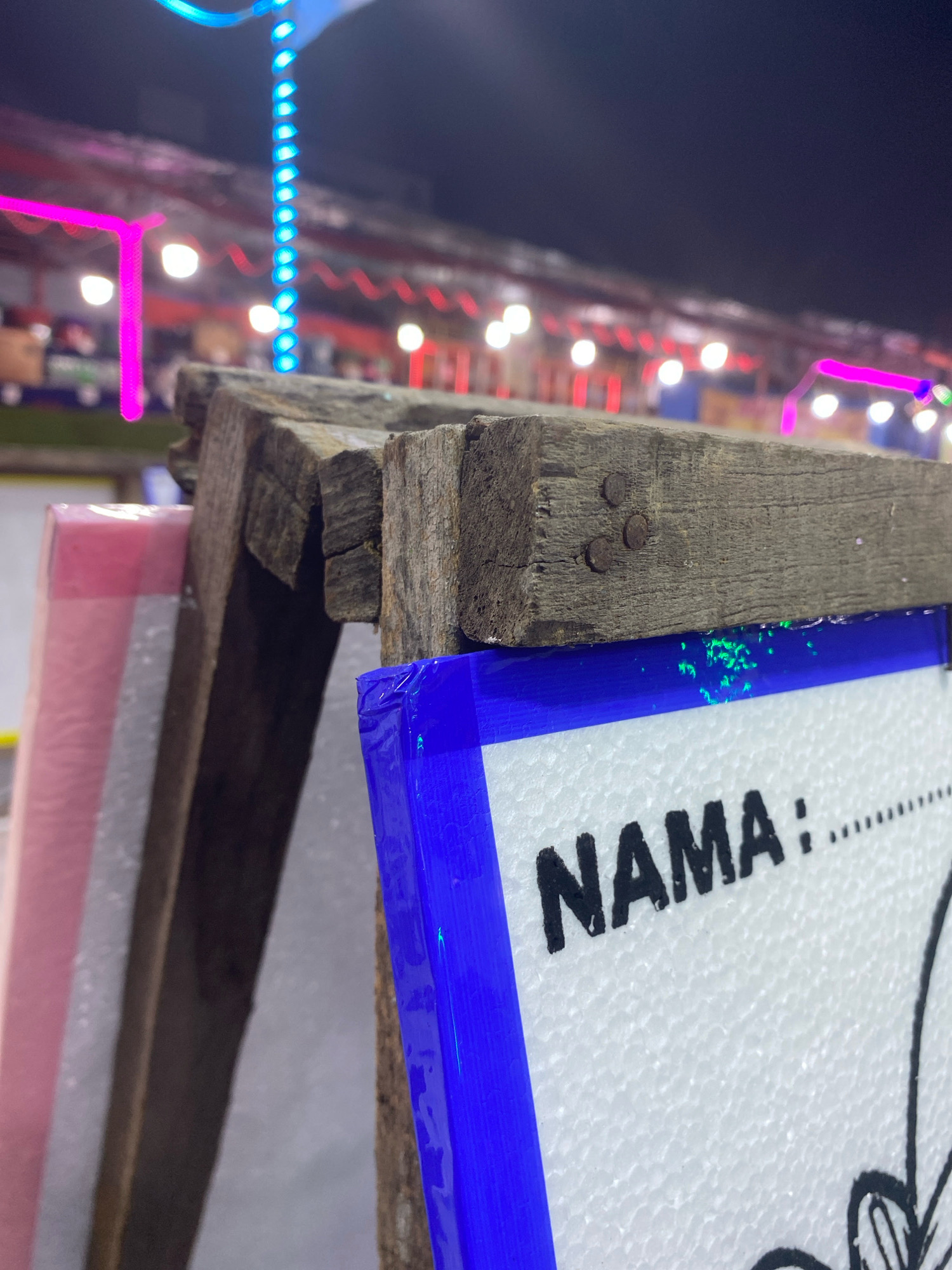 This image, taken at night in Rejosari, Indonesia, shows a close-up view of a rustic wooden frame holding a white sign with the word "NAMA" printed in bold black letters, followed by a dotted line. The sign is bordered with a bright blue tape, with some green light reflecting on the top edge. Behind the sign and frame, out-of-focus pink and white objects are visible. In the background, blurred colorful lights, including blue and pink strands, illuminate what appears to be a festive or recreational area, possibly a market or an outdoor event. The overall impression is one of a vibrant nighttime setting, with the foreground focusing on an instructional or identification element.