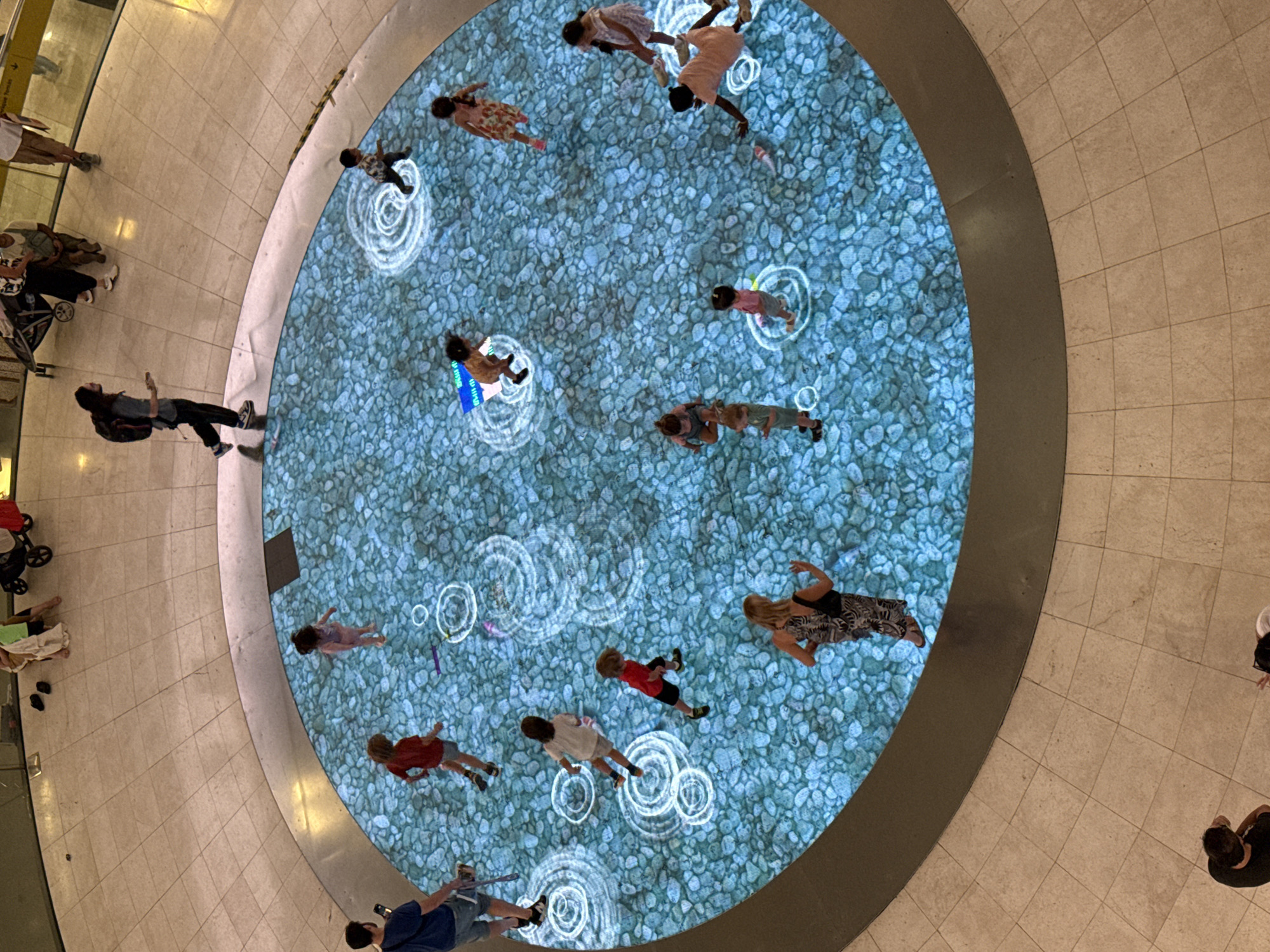 In Barcelona, Spain, a circular interactive water feature is located in an indoor public space, possibly a shopping mall or plaza, with beige tiled flooring. The water feature's surface displays a high-resolution projection of blue pebbles and animated ripples, creating the illusion of water. Several children are interacting with the projection, their footsteps triggering circular wave patterns.
From the top left, moving clockwise:
A man wearing a blue t-shirt and shorts stands on the tiled floor, holding a phone.
A child in a red shirt and shorts runs across the projection.
A child in a pink outfit stands on the projection.
A child in a patterned dress stands on the projection.
A child in a pink shirt and shorts is crouched on the projection.
A woman in a black and white patterned dress stands on the edge of the projection, looking into it.
A child in a light peach outfit is partially visible, reaching towards the projection.
A child in a green outfit stands on the projection.
A child in a patterned dress stands on the projection.
A child in a red shirt and black shorts runs on the projection.
A child in a light beige outfit is crouched on the projection.
A child in a white shirt and shorts runs on the projection.
A child in a patterned outfit runs on the projection.
A man with long hair, wearing a grey t-shirt, black pants, and a backpack, walks on the tiled floor.
In the background, other individuals and a stroller are visible on the tiled floor. The overall scene captures children engaged with an interactive digital display in a modern public environment.