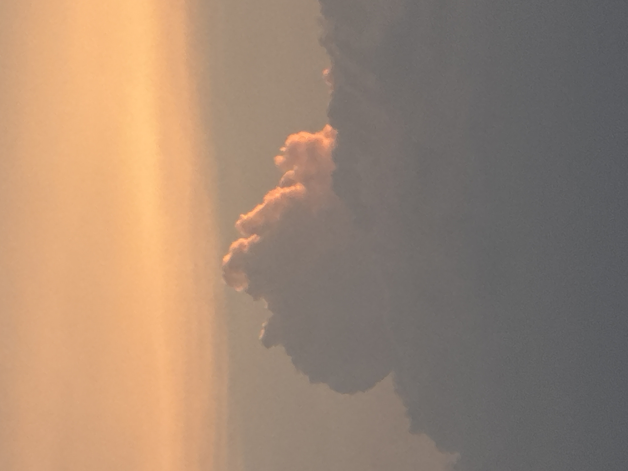 The image depicts a sky scene, likely at dusk or dawn, as indicated by the warm, golden light on the left side. This light illuminates a cluster of clouds, some of which are tinged with a pink hue. To the right, a larger mass of dark clouds dominates the view. The photo's orientation is vertical, suggesting it may have been taken with a mobile device. The specific location within San Francisco Ocotlán (Ocotlán), Mexico cannot be determined solely from the image. There is no further contextual information about people, subjects, objects, actions, interactions, or the environment.