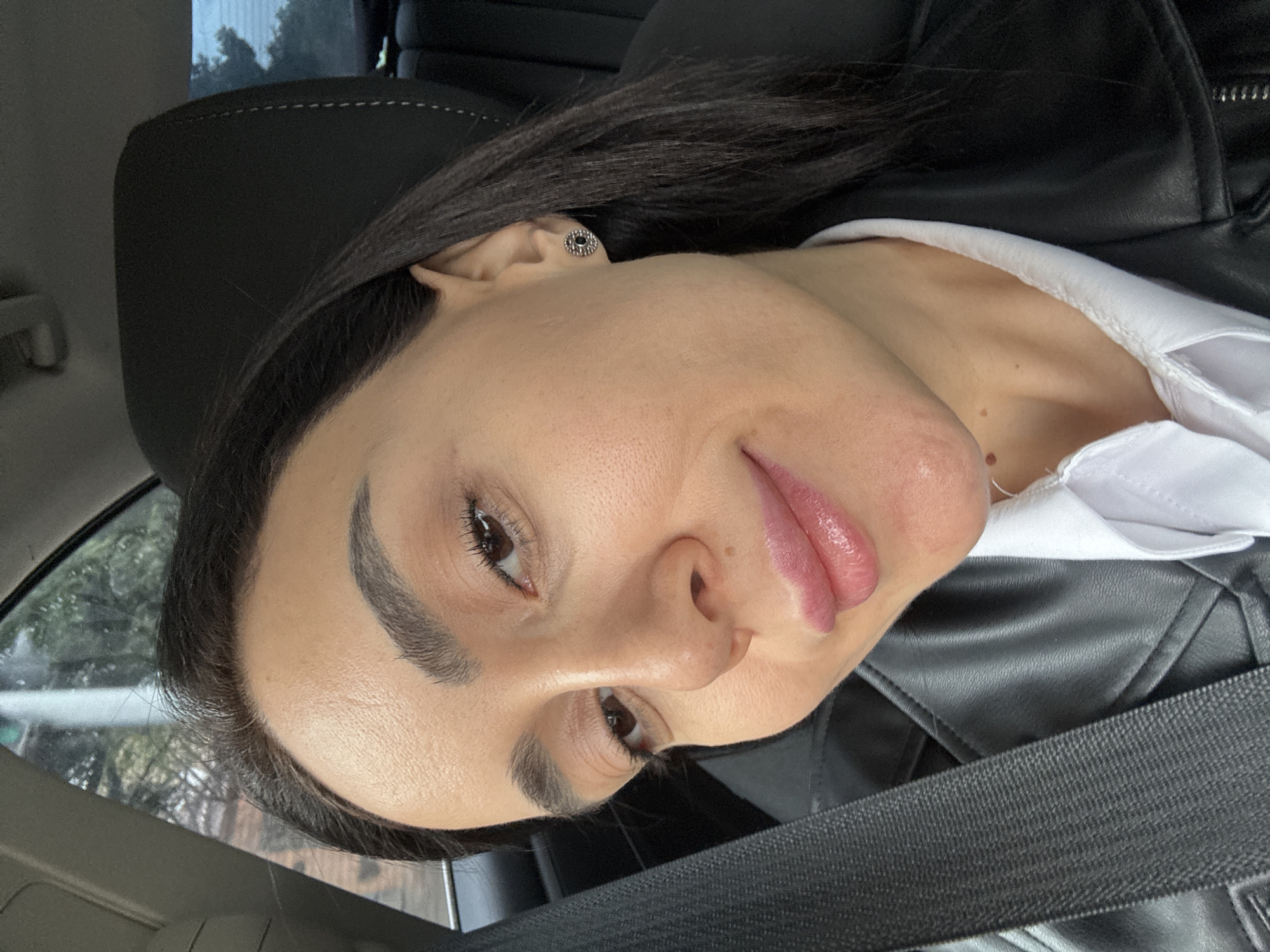A dark-haired woman is seated in a car, wearing a black leather jacket and a white shirt. She is secured with a seatbelt. Her head rests against the headrest of the car seat. She has fair skin, dark eyebrows, and dark eyes. Her lips are slightly parted. She is wearing a silver earring. The image was not taken in the city of Tbilisi, Georgia and no assumptions about the location can be made with this information.