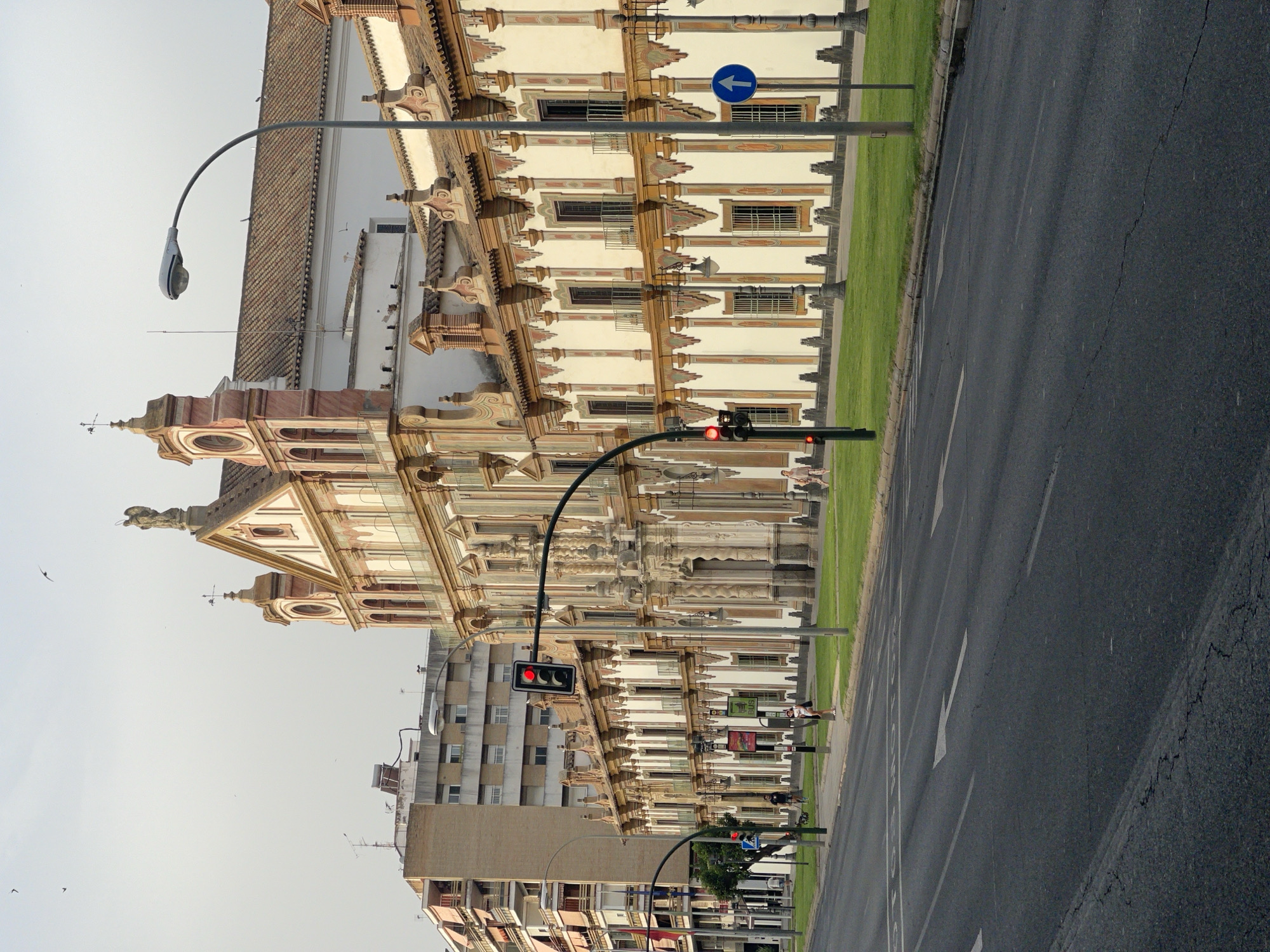The image depicts the Palacio de Congresos y Exposiciones (Congress and Exhibition Palace) in Córdoba, Spain. The foreground features a multi-lane asphalt road with visible lane markings and traffic signals. One traffic light is red, indicating a stop. A blue directional sign with a white arrow pointing upwards is visible on the right side of the road. To the right of the road, a neatly manicured green lawn borders the palatial building.
The building itself is a grand structure with a light-colored facade adorned with decorative elements in brown and reddish-pink. It features numerous windows with intricate detailing and small balconies. The architecture suggests a historical significance, with a prominent central entrance and ornate rooflines. A statue of an angel is visible atop the central gable.
Several people are present on the sidewalk and in the background. On the sidewalk in front of the building, a person is walking a dog, and another individual is standing. Further down the sidewalk, two women are walking away from the camera. In the background, a tall, modern apartment building stands to the left of the palace, contrasting with the older architecture. The sky is overcast with a few birds flying overhead. The overall scene captures an urban environment with a blend of historical and modern architecture.