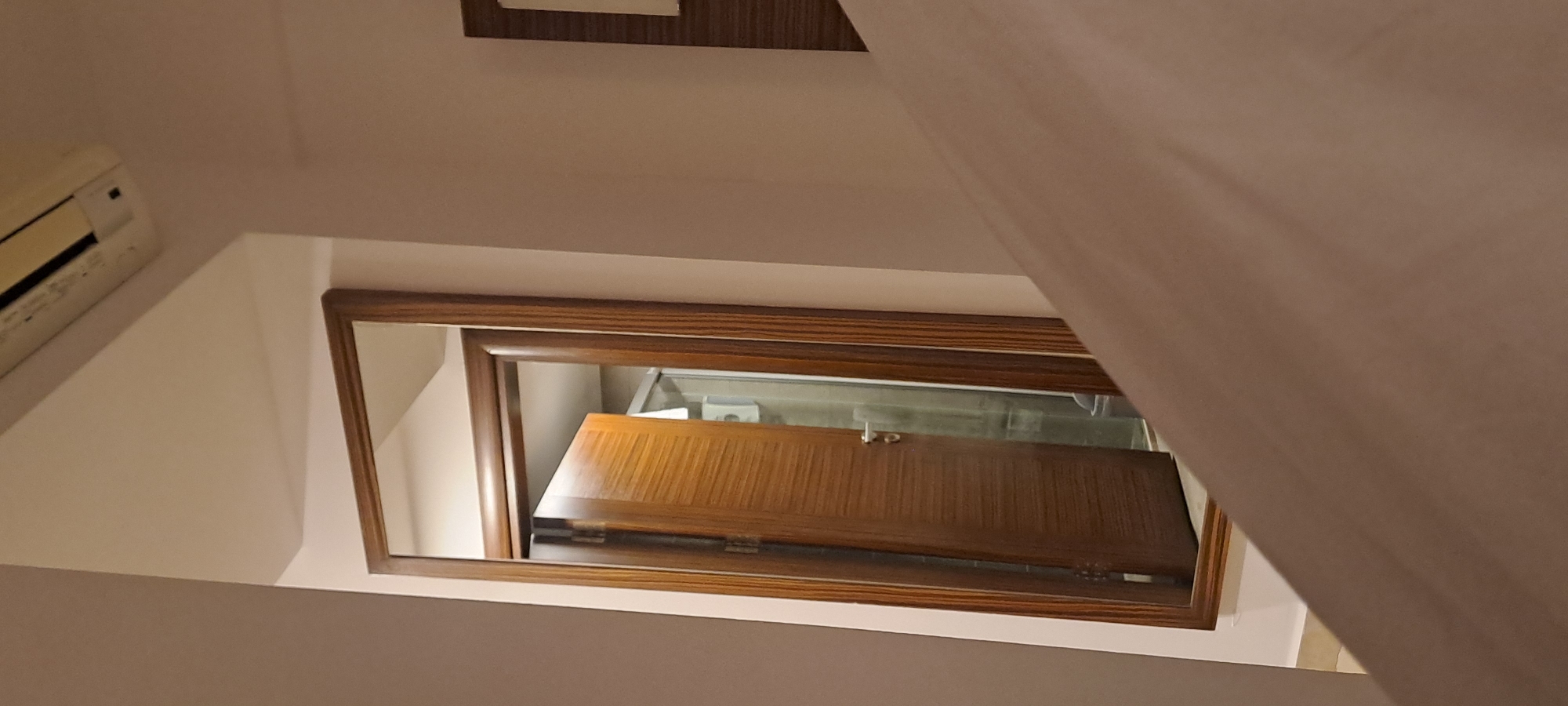 This image displays an interior room, identified as being in Kuşadası, Turkey. The primary subject is a large, rectangular mirror with a thick, dark wooden frame, mounted vertically on a light-colored wall. The mirror reflects an open wooden door, featuring horizontal paneling, which leads into an adjoining space. Within the reflection of the doorway, a glass shower enclosure with a silver handle, a white toilet, and a grey waste bin are discernible in the background. Above and to the right of the mirror, a portion of a white air conditioning unit is visible on the upper wall. The lower right foreground of the image is occupied by an out-of-focus area of beige bedding, suggesting the perspective is from a bed. The walls are a uniform light hue. No persons are present in the scene.