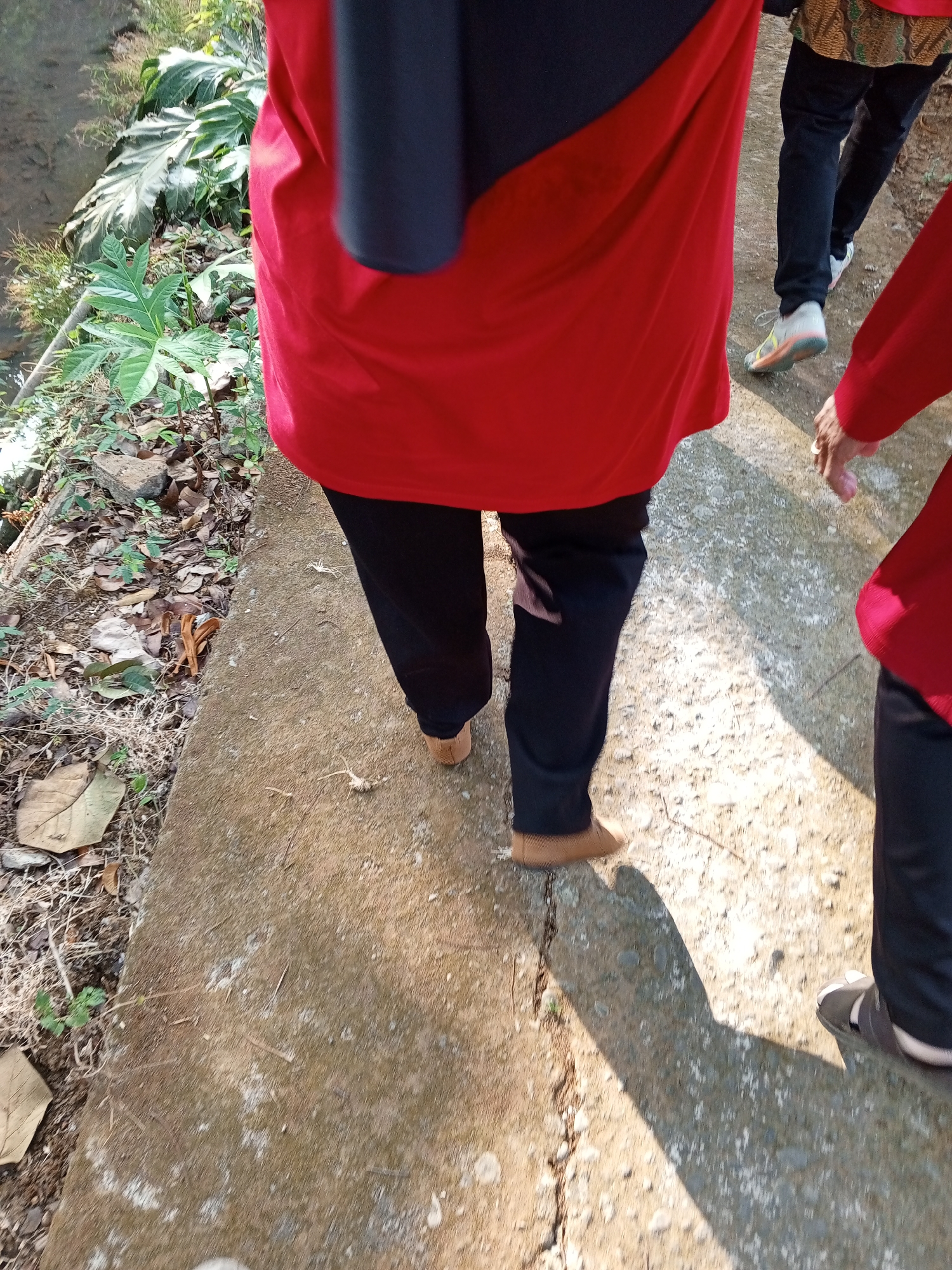 The image shows the lower bodies of at least three individuals walking on a concrete path next to a grassy area with some foliage. The path is cracked and uneven, with dirt and dry leaves on the ground beside it. The individuals are wearing casual clothing, including black pants and red tops. One individual on the left is wearing brown slip-on shoes, while another in the background is wearing white and colorful athletic shoes. The scene appears to be outdoors, possibly in a park or a rural area. The sunlight casts long shadows on the path, indicating it is daytime. The location is identified as Lemahbang, Indonesia, suggesting a tropical or subtropical environment given the lush vegetation visible. The presence of multiple people walking suggests a shared activity or simply passing by each other. The overall impression is one of a casual outdoor stroll in an Indonesian setting.