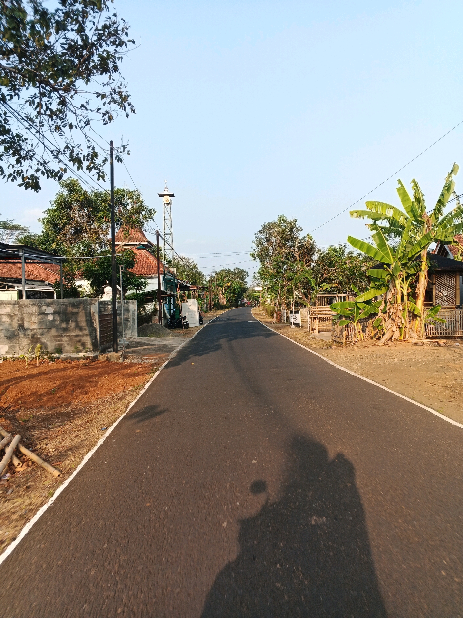 The image depicts a paved road in what appears to be a rural or semi-rural area of Pesanggrahan, Indonesia. The road is dark asphalt with a white painted line along the right edge, indicating the shoulder. The perspective suggests the photo was taken from a moving vehicle, likely a motorcycle, as indicated by the prominent shadow of a person and a motorcycle on the road surface.
On the left side of the road, there is a concrete wall and a fence, behind which are buildings with terracotta tile roofs and trees. A tall metal tower with a bell or speaker at the top is visible, possibly a call to prayer tower or a public announcement system. There are also power lines stretching across the scene. The ground to the left of the wall is covered in reddish-brown soil and some dry vegetation.
On the right side of the road, there are banana trees, a small bamboo structure that resembles a hut or shelter, and a wooden fence. More trees are scattered throughout the background. The sky is clear and blue, suggesting a sunny day. The overall impression is a quiet, roadside scene in an Indonesian village.