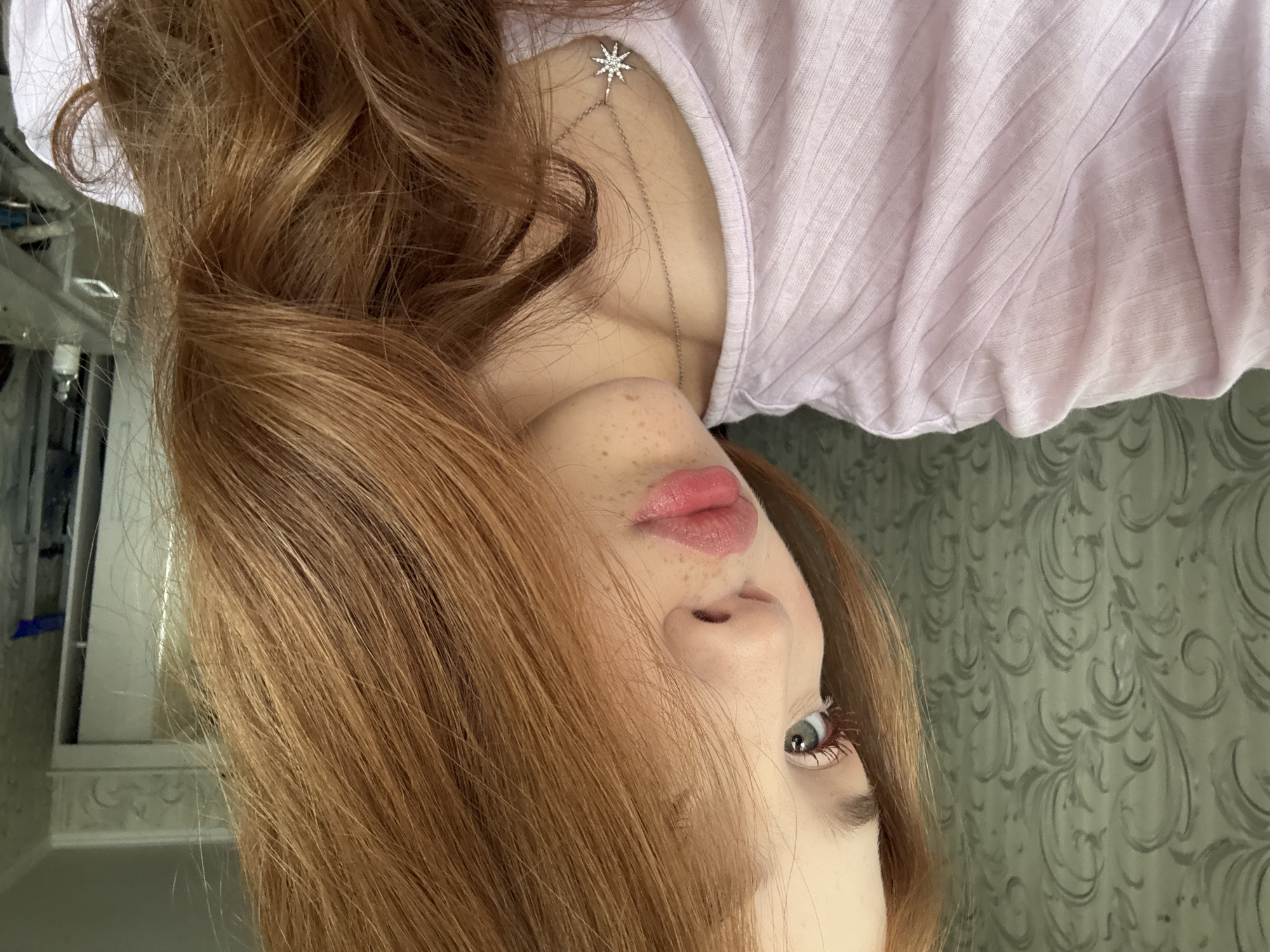 A young female subject is depicted in a close-up, side-profile portrait within an indoor residential setting, purported to be located in Naberezhnye Chelny, Russia. The subject has long, reddish-brown, wavy hair that partially obscures the right side of her face. Her left eye, light blue in color, is visible, along with defined eyelashes and a sculpted eyebrow. Freckles are discernible on her nose and upper lip area. Her lips are full, glossy, and tinted with a pink hue, adopting a slight pout. She wears a light pink, ribbed garment. A silver chain necklace with a star-shaped pendant, embellished with small reflective stones, rests on her upper chest. The background on the left features a wall covered with light green patterned wallpaper featuring swirling motifs. On the right, an indistinct background includes a white door frame or mirror and faint outlines of other room elements such as a surface, bottles, and a blue object.