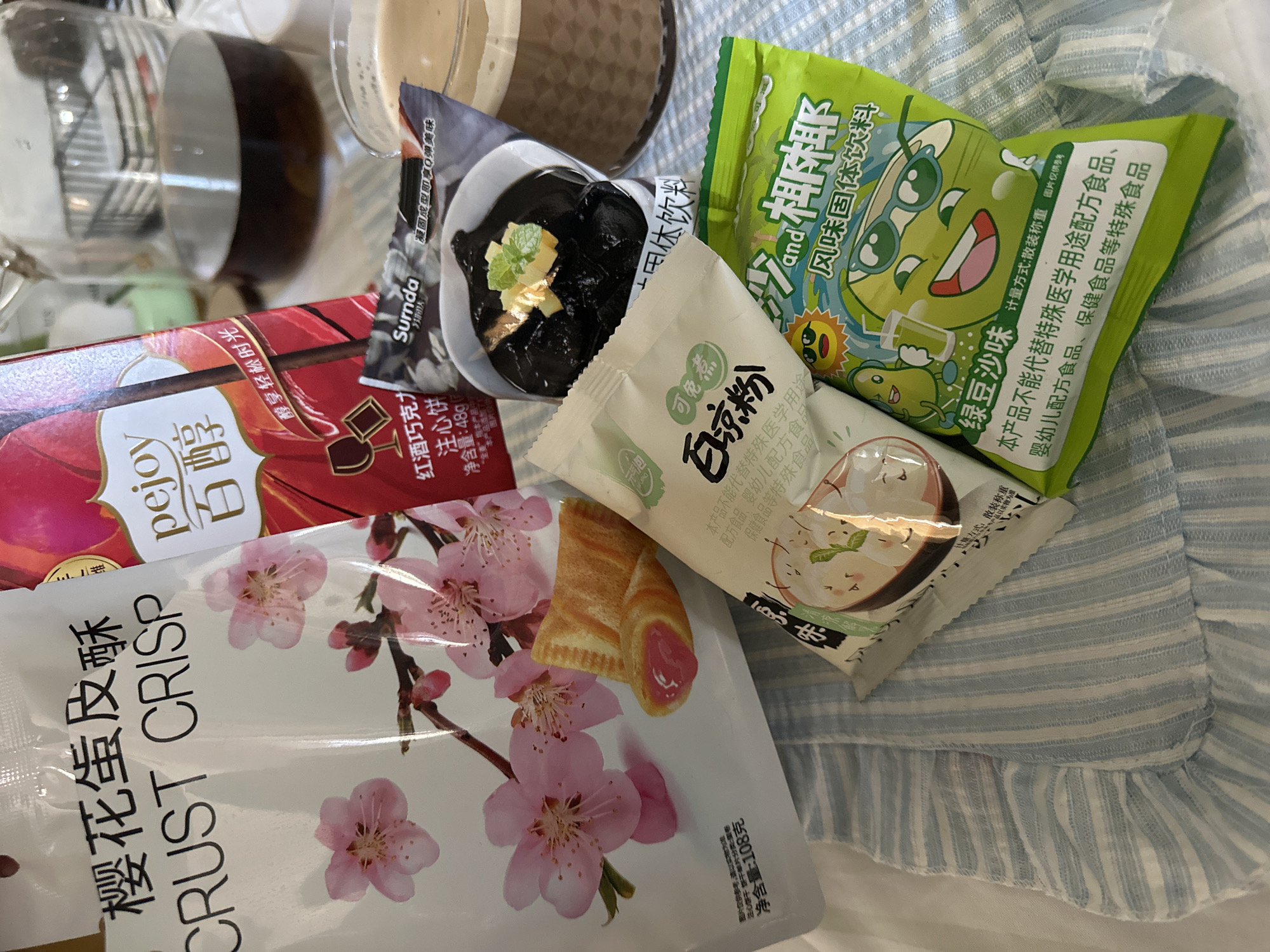 The image shows a collection of packaged food items on a blue and white striped surface with a ruffled edge. The food items include: "CRUST CRISP" pastry with cherry blossom imagery, "pejoy" chocolate sticks, "Sumda" jelly dessert, "Bai Liang Fen" powder, and a green packet of "Lv Dou Sha Wei" bean-based drink. The scene is indoors and objects are arranged haphazardly. The image does not suggest a specific location in Bar, Montenegro.