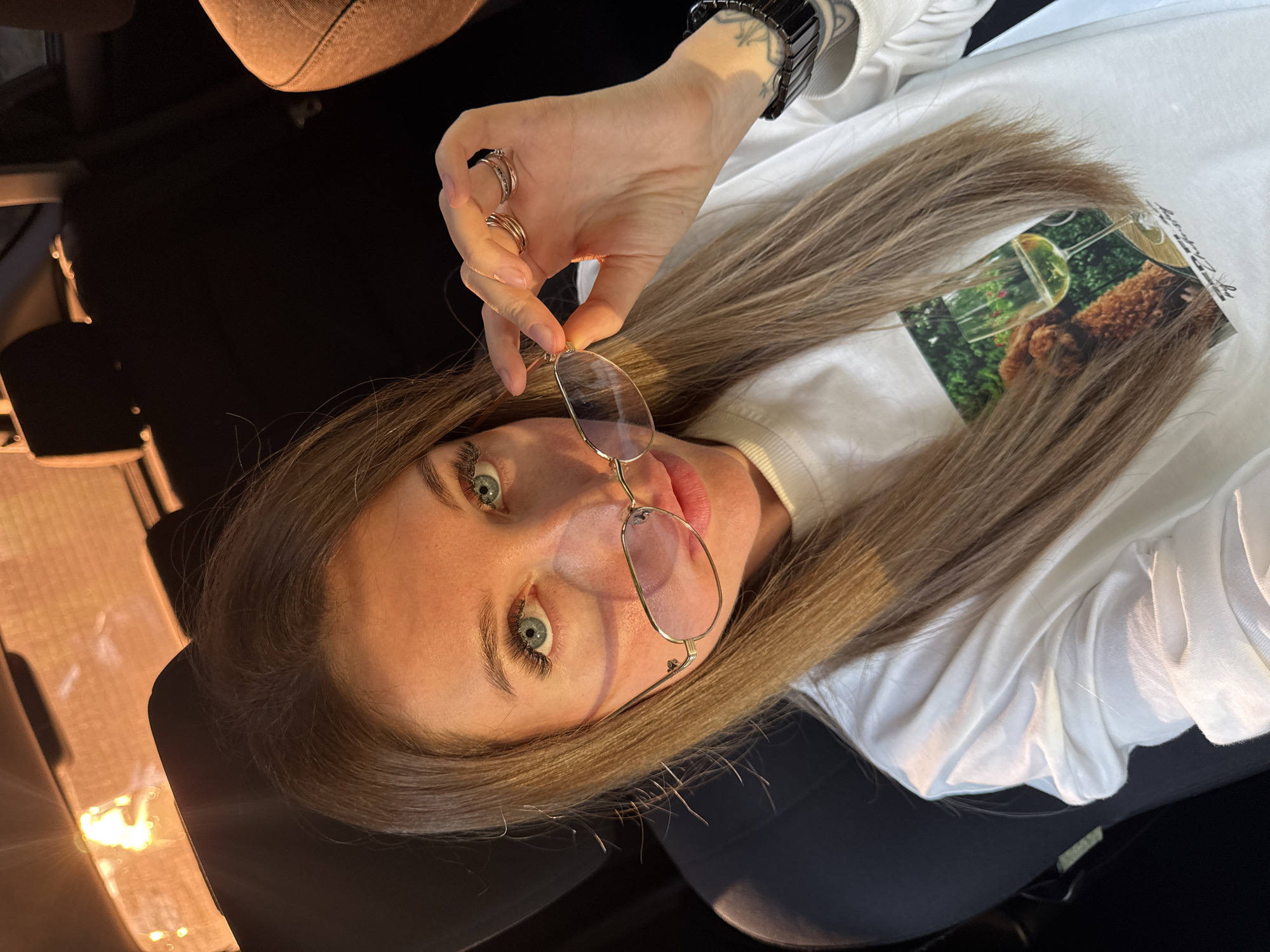 A woman with blonde hair and blue eyes is sitting in a car, holding her glasses. She is wearing a white sweatshirt with a picture of a dog and a glass of wine on it. The car interior is dark, with the sun shining through the windshield. The image is taken from a high angle, looking down at the woman. There is no identifiable information about the location being in Chaikovsky, Russia.