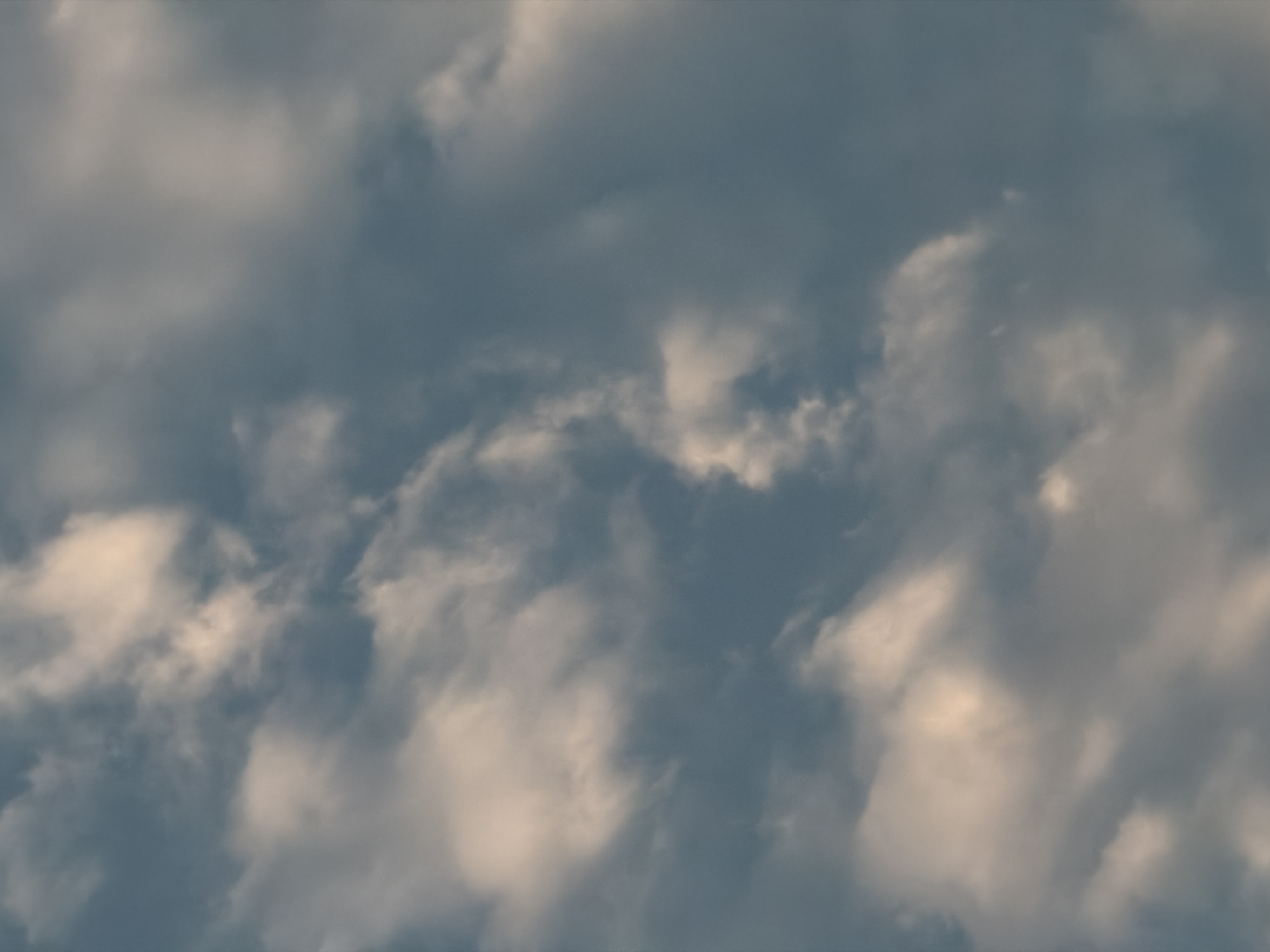 The image captures a detailed, abstract view of a cloudy sky over Hải Phòng, Vietnam. The scene is filled with a dense array of clouds, exhibiting a varied palette of grey, blue-grey, and pale yellow-white tones. Sections of the clouds appear brightly illuminated, suggesting sunlight is breaking through or reflecting off their surfaces, creating soft, warm highlights, particularly on the edges and upper parts of some formations. These brighter areas contrast with the deeper grey and blue-grey masses, which convey a sense of depth and density. The textures range from soft, wispy veils to more substantial, billowy structures. There are no discernible objects, people, or identifiable landmarks within the frame; the focus is solely on the intricate patterns and atmospheric conditions of the sky. The interplay of light and shadow on the clouds suggests a dynamic weather pattern, possibly an overcast or partly cloudy day, or a specific time of day such as sunrise or sunset where light filters through atmospheric layers. No activity is depicted, and no text is visible.