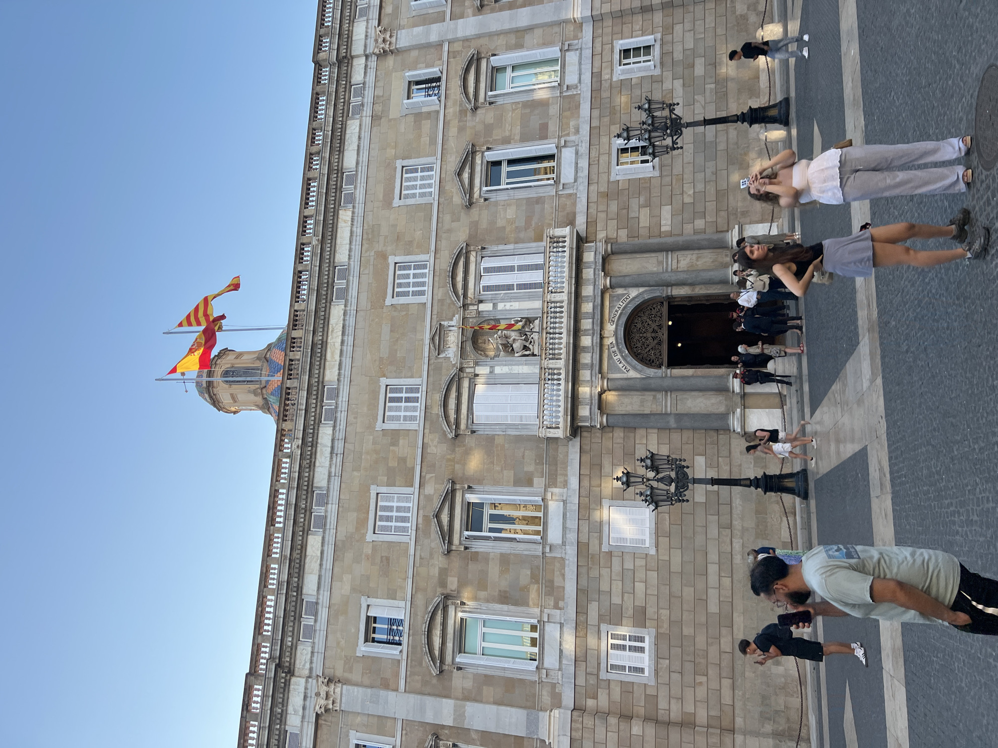 This image shows the Palau de la Generalitat de Catalunya, a historic building located in Barcelona, Spain. The building features a stone facade with numerous windows, some of which are open, revealing white shutters. Two flags, the Spanish flag and the Catalan flag, fly from poles atop a dome on the building. In front of the building, several people are visible. Two women in the foreground are taking photos with their phones. Other individuals are standing or walking in the plaza, including a man holding a phone, and a police officer near an entrance. The ground in front of the building is paved with a patterned stone. The sky is clear and blue.