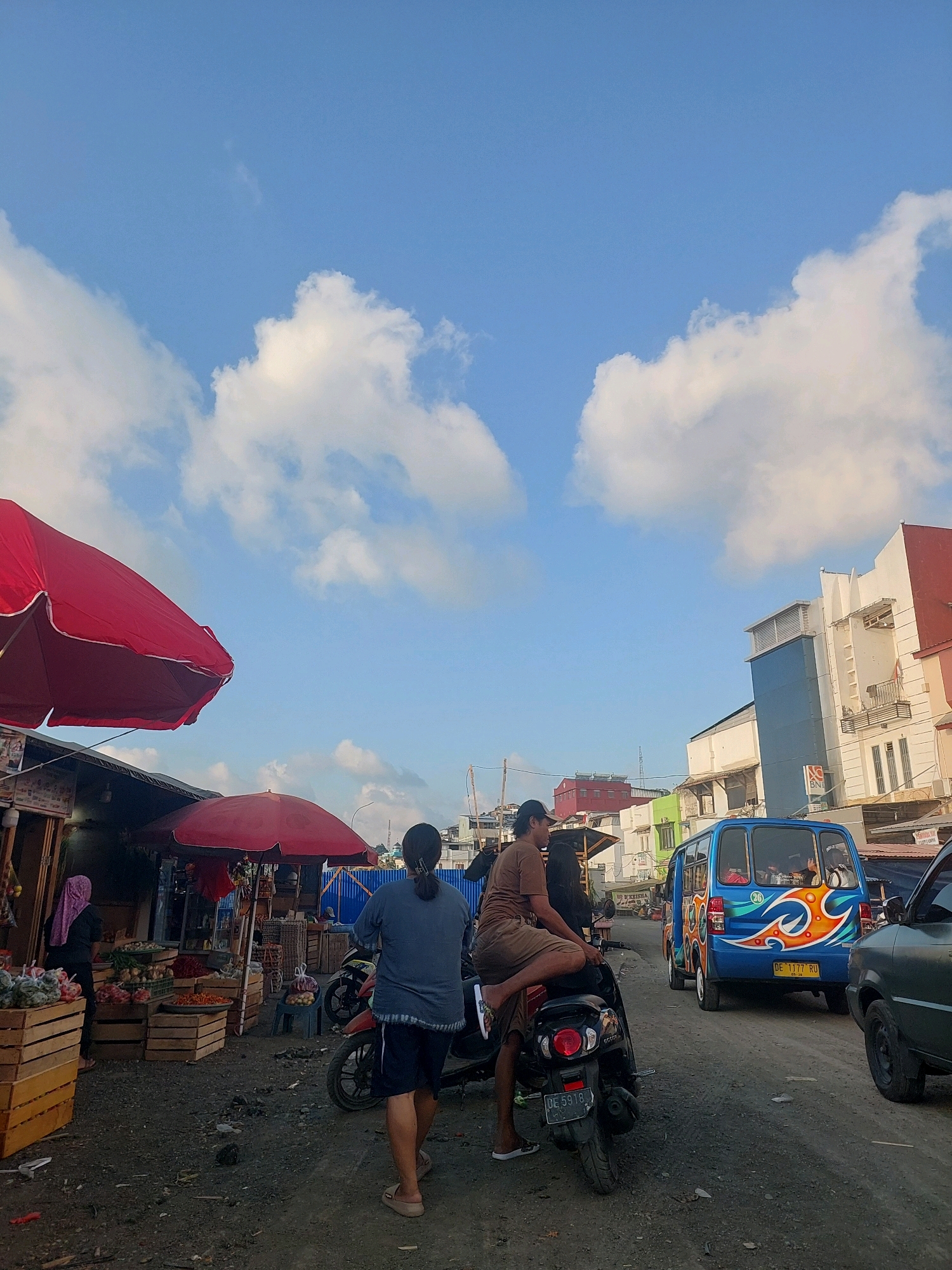 The image captures a street scene in Ambon, Indonesia, likely a market area. The sky is blue with scattered white clouds. In the foreground, two individuals are on a motorcycle, one driving and the other seated behind, facing left. To the left, a market stall is visible with red umbrellas, displaying produce in wooden crates, and a person in a pink headscarf. Further down the street, a colorful blue minibus with orange and white accents is driving away, followed by a dark green car. Several buildings line the right side of the street, some appearing to be commercial establishments. The ground is unpaved and appears dusty.
