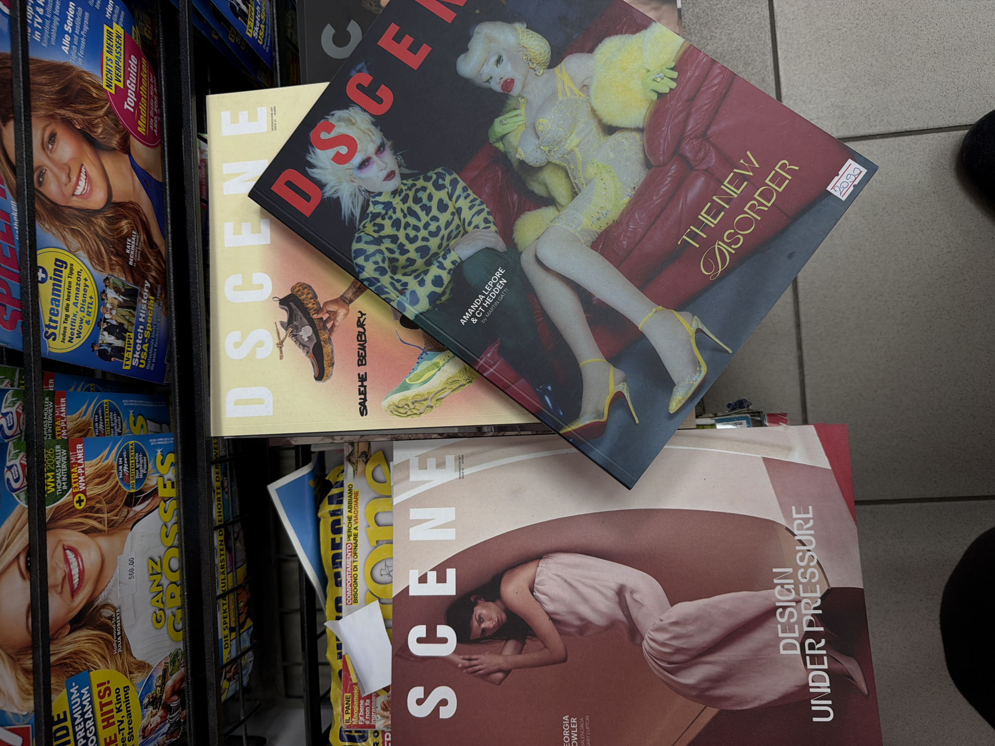 The image shows a magazine rack filled with various magazines. The topmost magazines are "DSCENE," with "THE NEW DISORDER" on one cover, and a second magazine "SCENE" with "DESIGN UNDER PRESSURE" on the cover. Other magazines are visible in the rack below, with various cover images and titles in German and Italian. The image appears to be taken at a magazine stand. The location of the image is not discernible.