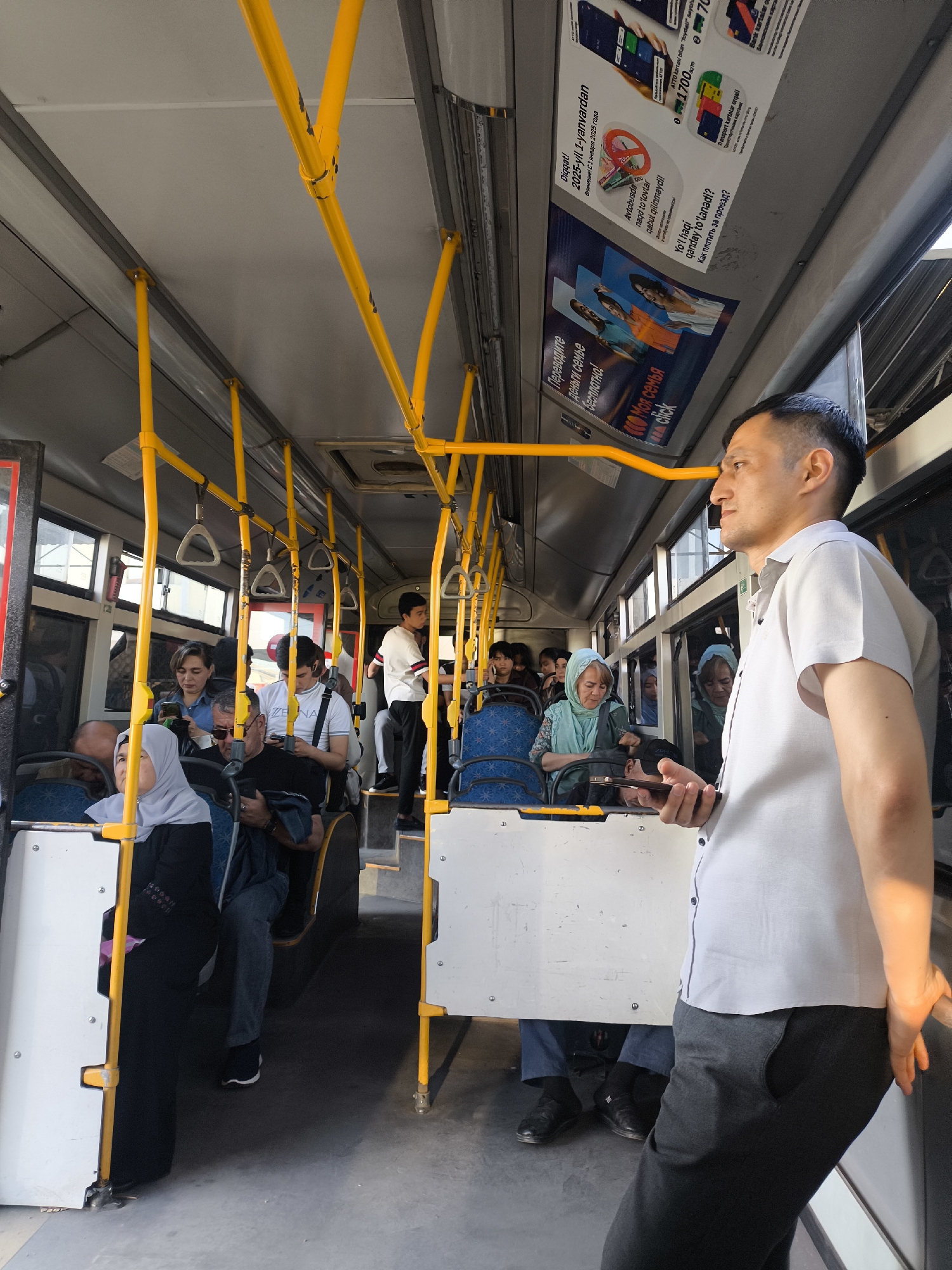 The image shows the interior of a public bus in Tashkent, Uzbekistan, filled with passengers. The bus has yellow handrails and advertisements displayed above the windows. Passengers are seated and standing, some looking at their phones. An Asian man in a short-sleeved white shirt and dark pants is standing near the front of the bus, looking out the window. Some of the passengers are women wearing headscarves. The bus appears to be in motion.