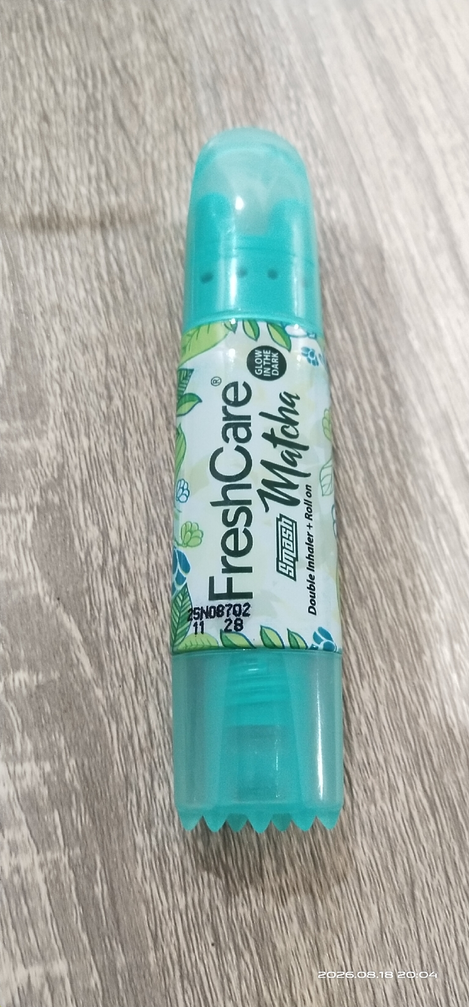 A teal-colored inhaler with a clear cap sits on a wooden surface. The inhaler has a label with a white background and a green leaf pattern. The label reads "FreshCare Matcha" in green text. Below "FreshCare", there is a smaller oval shape with "GLOW IN THE DARK" written vertically. Below "Matcha", it says "Double Inhaler + Roll on". There is also some text and numbers in black: "25N08702 11 28". The bottom of the inhaler has a teal plastic cap with a serrated edge. A timestamp in the bottom right corner of the image reads "2026.08.18 20:04". The image location is not specified in the provided information.