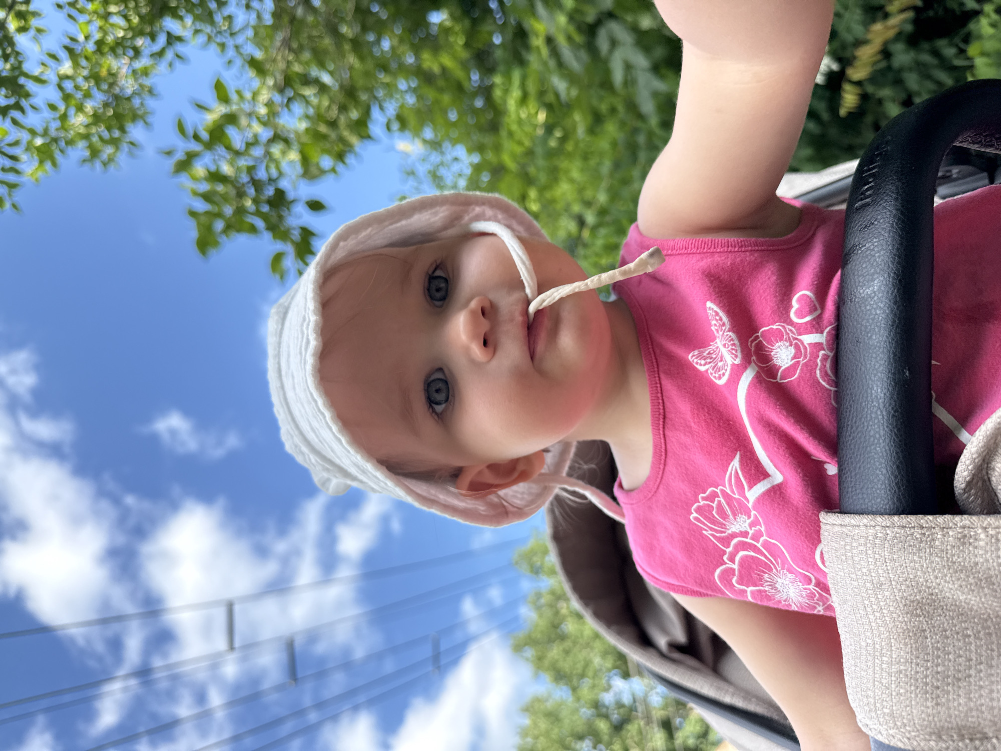 This is a close-up, vertically oriented photograph of a baby wearing a white sun hat and a pink tank top with a floral design. The baby is looking directly at the camera with a neutral expression. A string from the sun hat is in the baby's mouth. The background consists of a bright blue sky with scattered white clouds and blurred green foliage, suggesting an outdoor setting on a sunny day. A black stroller handle is visible on the right side of the frame. The image appears to be taken from a low angle, looking up at the baby. There is no visible text in the image. The scene is likely taking place during the daytime.