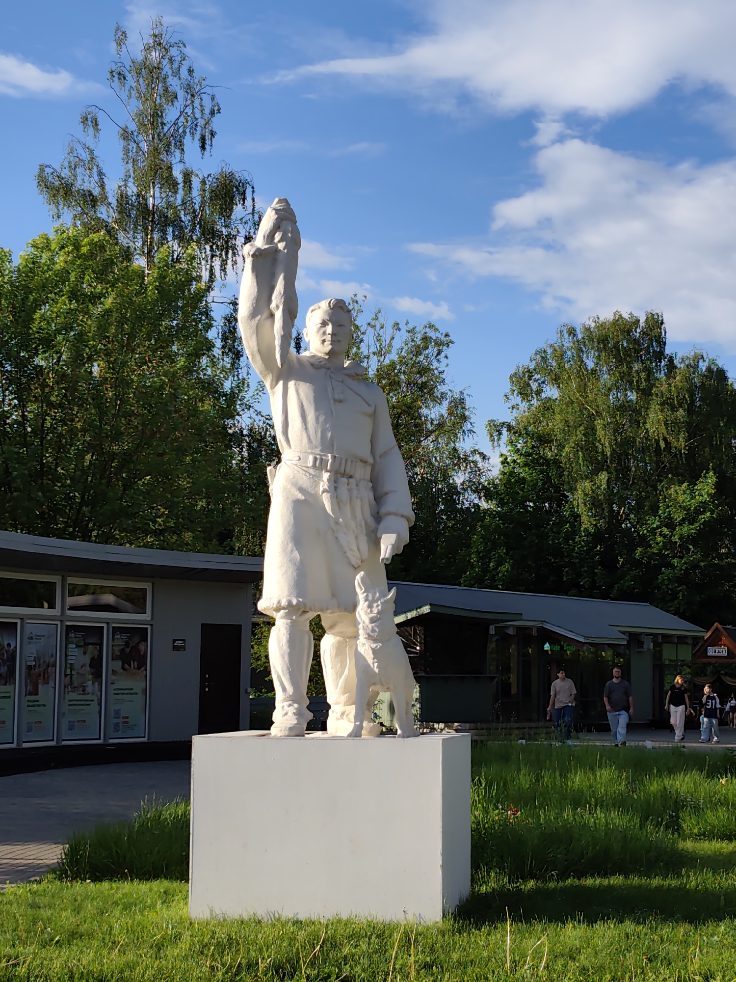 A full shot captures a white statue on a white pedestal, centered in the frame. The statue depicts a man holding a fish above his head, with a dog standing near his legs. Behind the statue are two low-rise buildings, with people walking on a pathway in the background. The sky is blue with scattered clouds, and green trees surround the scene. The location is likely a park in Moscow, Russia.