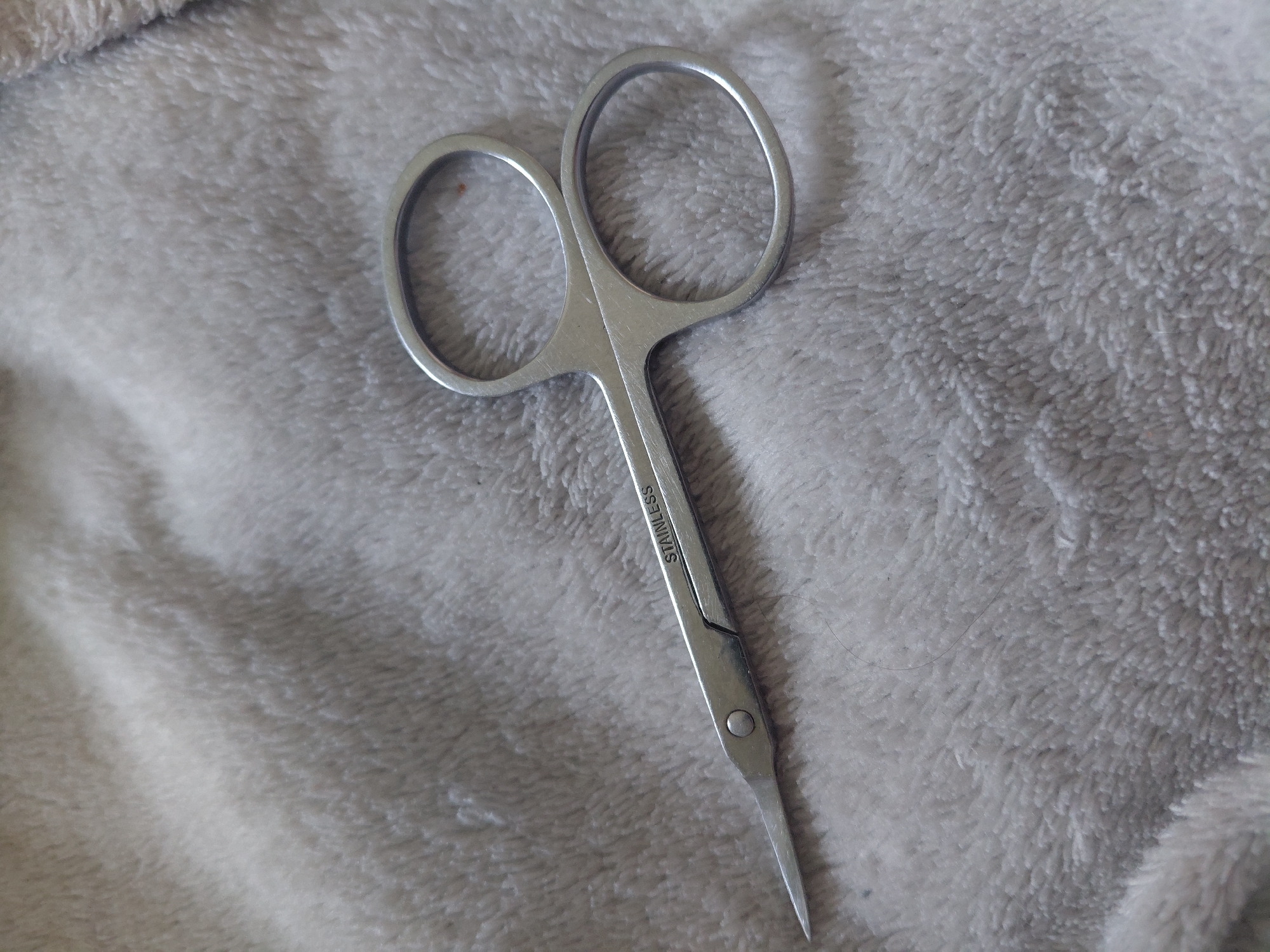 The image displays a pair of small, metallic scissors resting diagonally on a soft, light grey, textured fabric surface. The scissors are constructed from a silver-colored metal with a brushed or matte finish. The word "STAINLESS" is engraved in capital letters along the upper blade arm. The scissors feature two curved finger loops and slender, pointed blades, indicative of manicure or detail-oriented cutting tools. A small, dark, thin strand, consistent with a human hair, is visible on the fabric surface to the right of the scissor blades. The fabric appears plush or fleecy, exhibiting subtle variations in texture and light reflection, characteristic of a soft pile material. The scene is illuminated by soft, diffused light, creating gentle highlights and shadows that emphasize the fabric's texture. No individuals are present, and no actions or interactions are depicted. The observable scene is situated in the city of Kyiv, Ukraine.