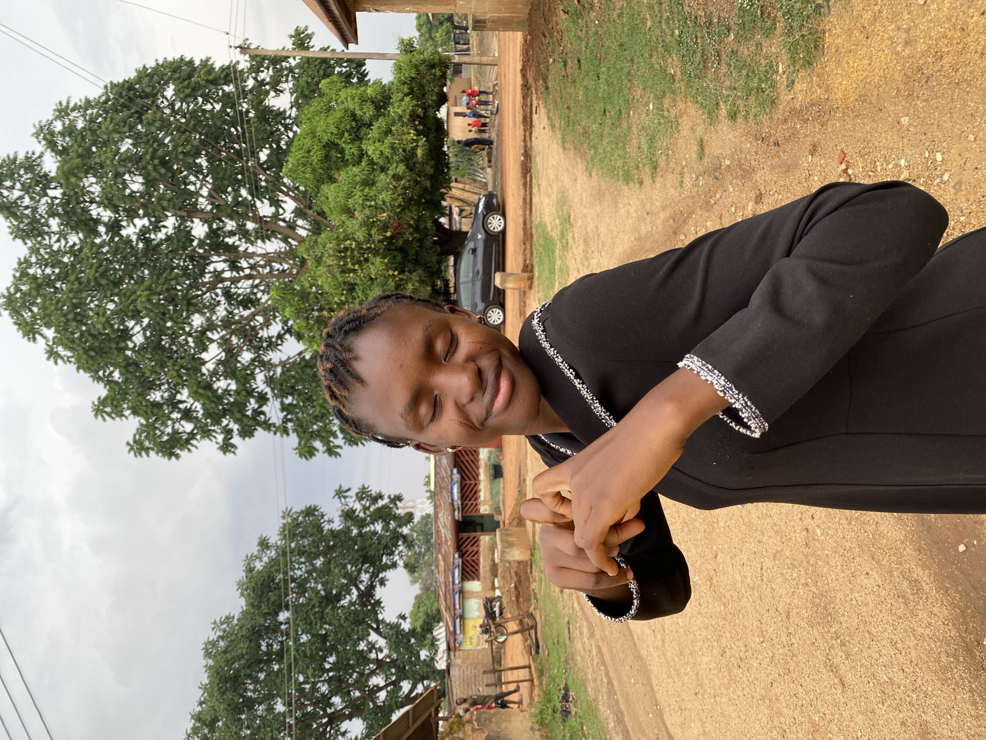 A young woman with braided hair is standing outdoors in Kuru Dungus, Nigeria. She is wearing a black garment with white trim and has her hands clasped. In the background, there is a dirt road, trees, buildings with corrugated metal roofs, a parked car, and people walking in the distance. The sky is overcast.