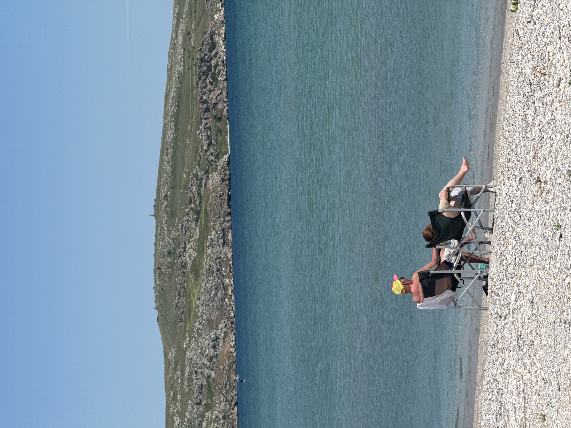 Two people are sitting in folding chairs on a beach near Мысовское сельское поселение, Ukraine. The beach is composed of small white stones. The water is calm and blue. To the left of the water, there is a rocky, green hill. The sky is clear and blue. The person closest to the camera is wearing a yellow hat, a black shirt, and black shorts. The other person is wearing a black shirt and patterned shorts.