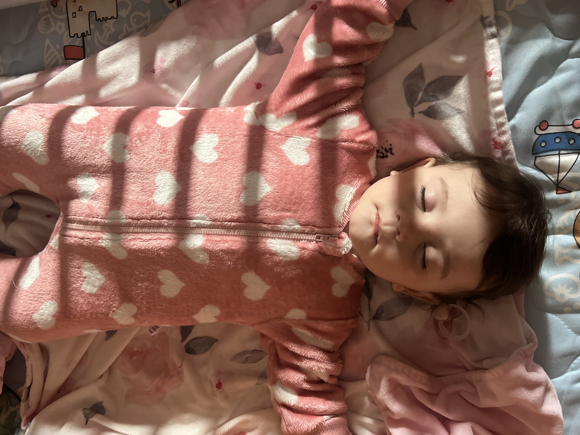 A baby sleeps supine on a bed. The baby wears a pink, long-sleeved onesie with white hearts. Shadows from a window or blinds fall across the baby's body. The bed has multiple blankets: a white blanket with pink floral designs, and a blue blanket with a drawing of a sailboat. There is no discernible information to indicate that the image was taken in Elena, Bulgaria.
