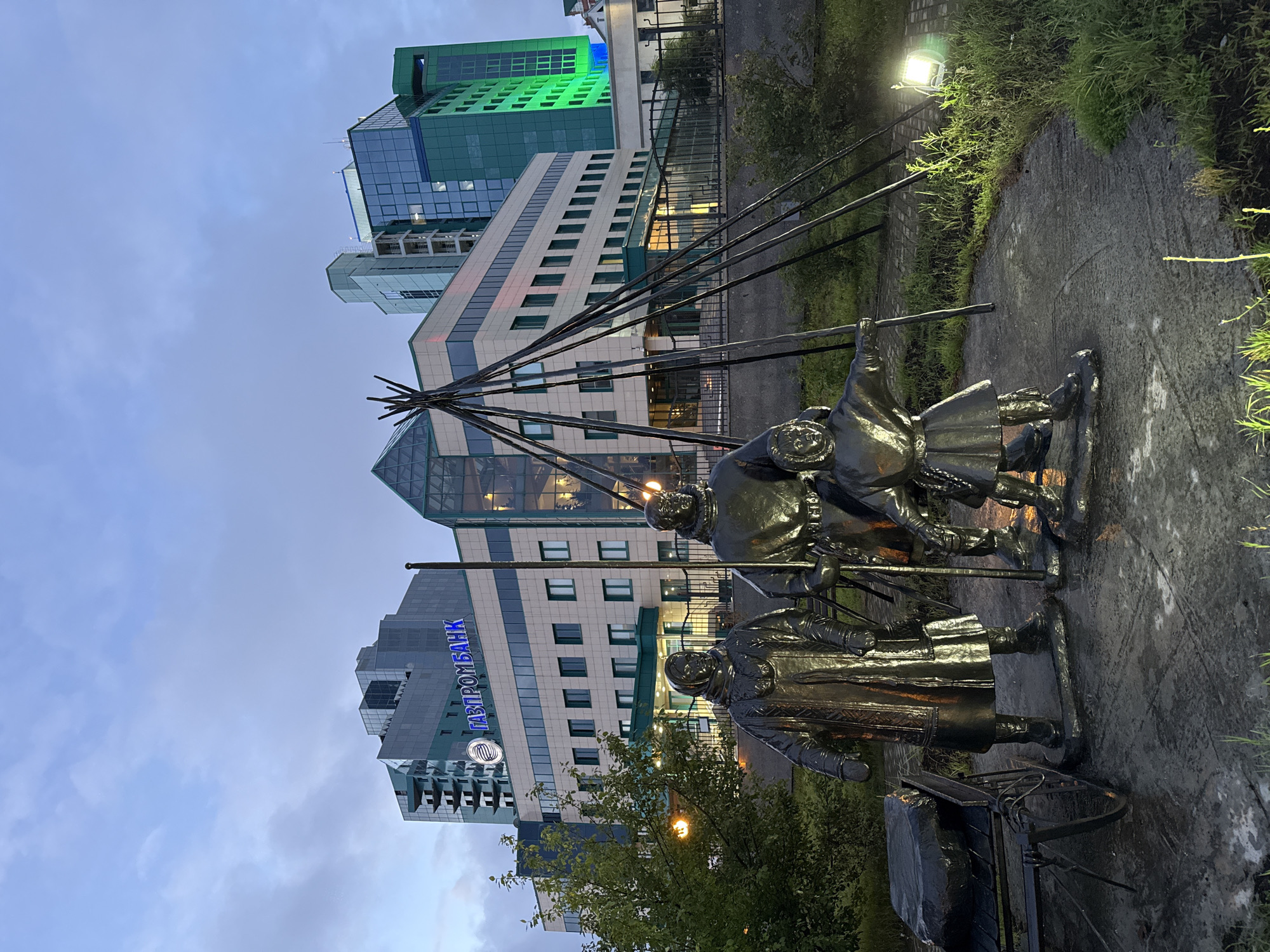 A bronze sculpture depicting three indigenous people stands in the foreground, with a modern office building in the background. The sculpture, which is wet from recent rain or dew, features two adults and a child dressed in traditional clothing. The adult on the left is wearing a long, fur-trimmed coat and a headscarf. The adult in the center holds a spear or staff, and the child on the right is reaching out a hand. To the left of the sculpture is a metal cart with a large, dark rectangular object on it.
The office building behind the sculpture is modern, with a glass facade and a prominent blue sign that reads "ГАЗПРОМБАНК" (Gazprombank). The building is illuminated, and the sky is overcast and appears to be twilight. The scene is set in Surgut, Russia, a city known for its oil and gas industry. The presence of the "Gazprombank" sign confirms the location as it is a major Russian bank. The overall impression is a juxtaposition of indigenous culture and modern urban development.