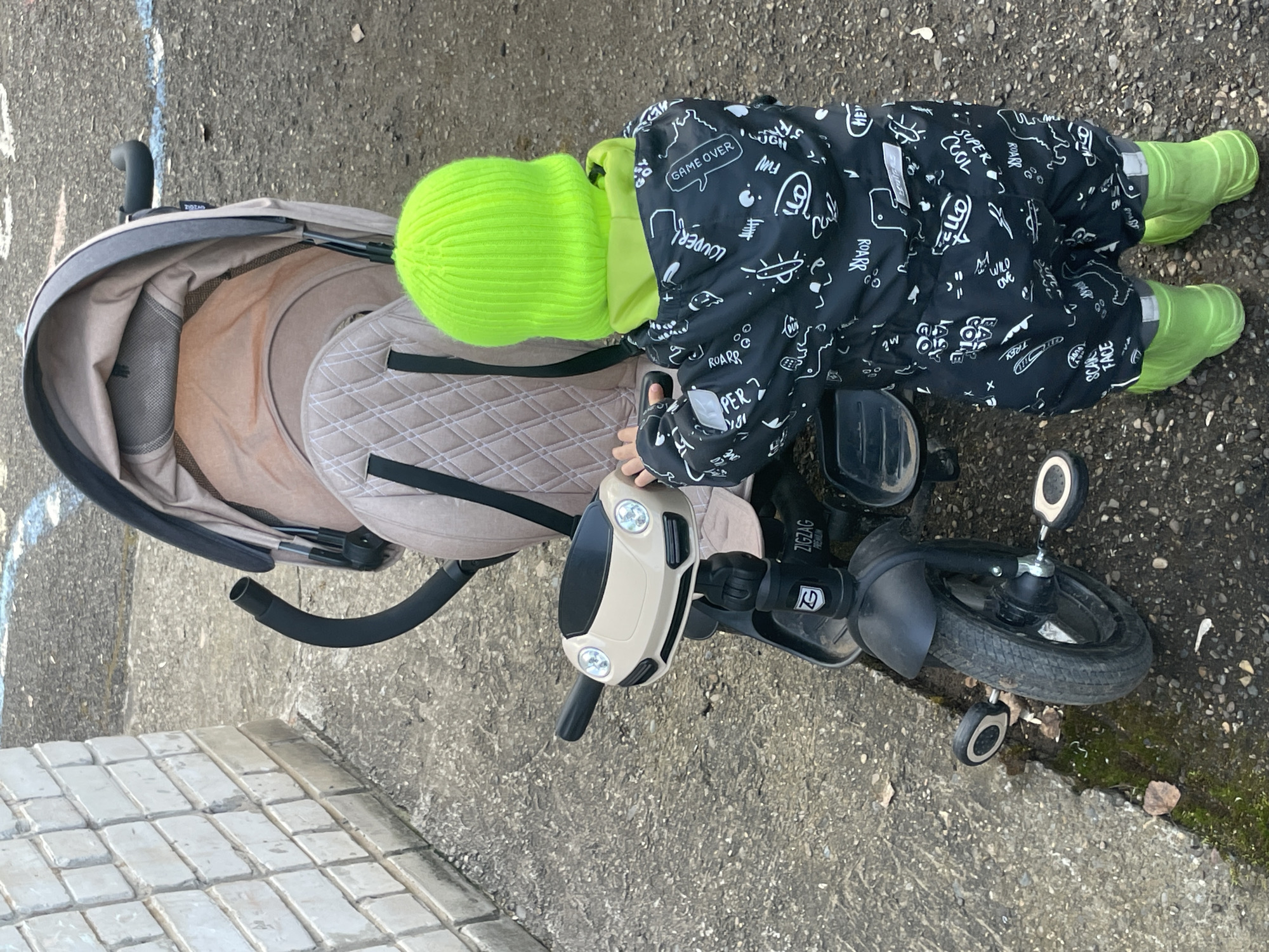 An overhead shot captures a young child, dressed for cool weather, engaging with a stroller and a tricycle on an outdoor concrete surface in Izhevsk, Russia. The child wears a bright neon green knitted beanie, a black and white patterned jumpsuit or snowsuit, and matching neon green rubber boots. The jumpsuit is covered with various cartoonish illustrations and text snippets including "GAME OVER," "LOUDER," "ROARR," "SUPER COOL," "WILD ONE," and "SCARY FACE."
The child is positioned between a beige stroller, which has a quilted diamond pattern on its interior, and a black tricycle-like ride-on toy with a beige steering mechanism designed to resemble a car's dashboard, complete with two round lights. The child's hands are near the handlebars of the tricycle. The stroller, visible on the left, has "ZIGZAG" visible on its frame. The tricycle also has "ZIGZAG PREMIUM" and a 'G' logo on its structure.
The ground is primarily rough gray concrete, with some lighter-colored rectangular paving stones visible on the left edge. Patches of moss and dried leaves are scattered on the right side of the frame, suggesting an outdoor, possibly urban or suburban setting. The overcast lighting indicates daytime without direct sunlight.