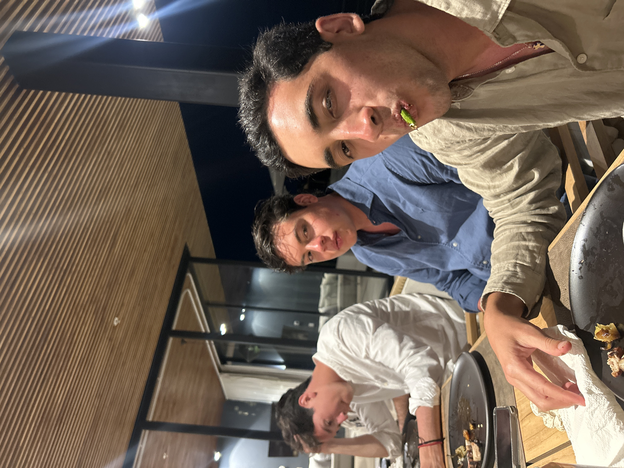 Three young men are at a dining table inside what appears to be a restaurant or upscale indoor/outdoor space. The location is speculated to be Cabo San Lucas, Mexico. One man is in the foreground with a green item in his mouth, wearing a light-colored shirt and a chain necklace. Another man is behind him, wearing a blue shirt. The third man is further back, wearing a white shirt and leaning towards the table. There are plates of leftover food and cutlery on the table, along with a white napkin being held. The interior features wood paneling and a dark sky is visible through the windows. There are lights above. The image has been rotated.