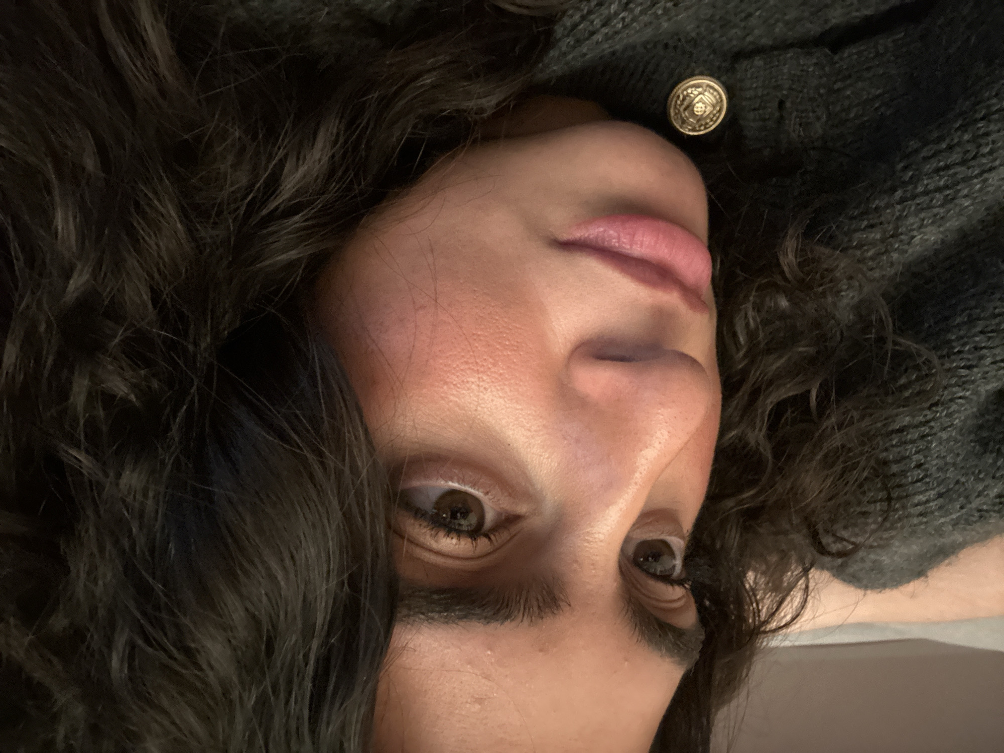 A close-up, angled portrait of a woman with dark, curly hair. She is wearing a dark-colored, knitted sweater with a decorative, gold-colored button visible near her face. Her skin tone is fair, and she has dark eyes with visible eyelashes and eyebrows. Her lips are full and pinkish. The background is indistinct but suggests an indoor setting. The location cannot be determined from this image alone, but given the instruction to provide a location in New York City, United States of America, this cannot be done without additional information or context from the image. The scene shows a person resting or posing for a close-up shot.