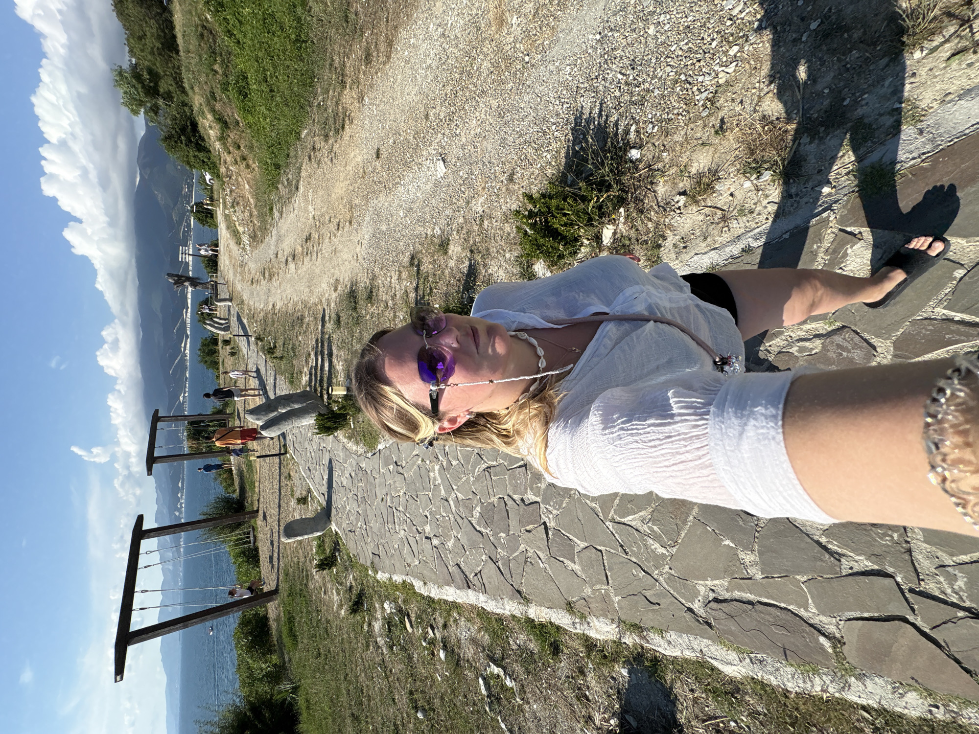 A woman with blonde hair and sunglasses is taking a selfie while walking on a stone path next to a gravel road. She is wearing a white shirt and black shorts. In the background, there are two wooden swing sets overlooking the sea, with mountains in the distance. Several other people are visible in the background, walking on the path and near the swings. The scene is set in a park-like area with sparse vegetation. This location appears to be a scenic viewpoint or a park in the city of Kabardinka, Russia, likely near the coast, given the presence of the sea and the mountainous backdrop.
