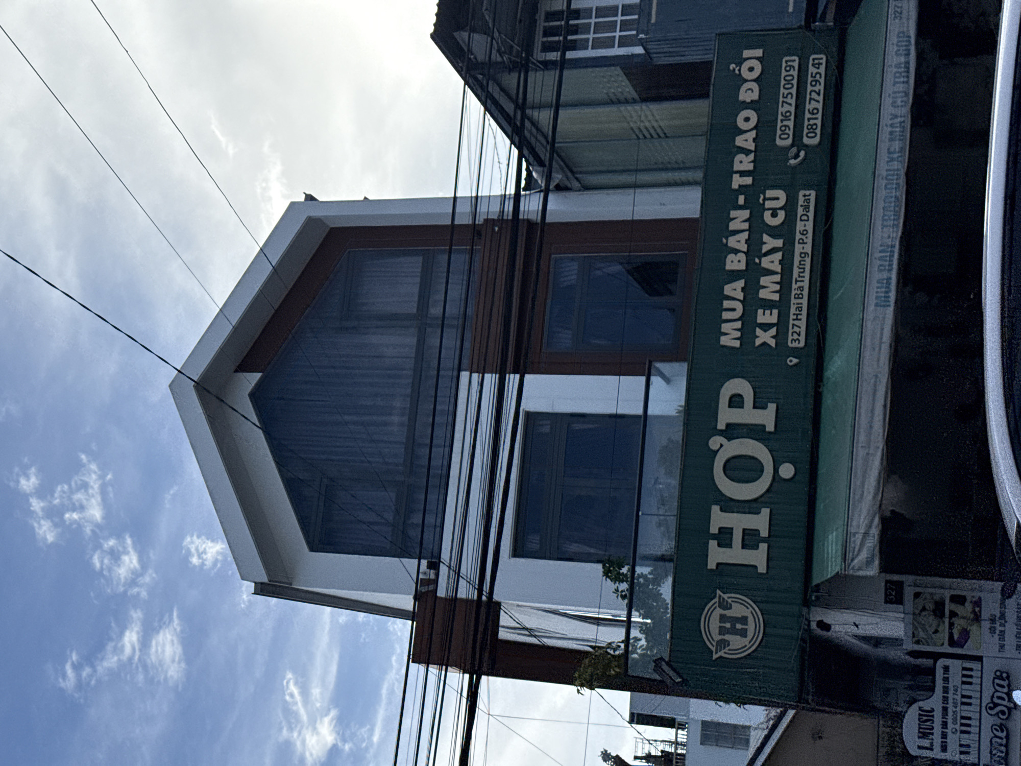 The image shows a modern, multi-story building with a distinctive angular design. The facade is a mix of white and dark brown, with large windows. Above the ground floor, a green sign displays the text "H OP" in large white letters, with a smaller wing logo to the left of the "H". Below this, a second green sign has white lettering indicating "MUA BÁN - TRAO ĐỔI XE MÁY CŨ" (meaning "Buy - Trade - Used Motorcycles"), an address "327 Hai Bà Trưng- P.6-Dalatt", and two phone numbers: "0916 750091" and "0816 729541". There are also smaller signs visible on the building. The sky is partly cloudy with a blue hue. Power lines are strung across the top of the image. A portion of a car is visible at the bottom. The scene appears to be a street in Da Lat, Vietnam, likely a commercial area.