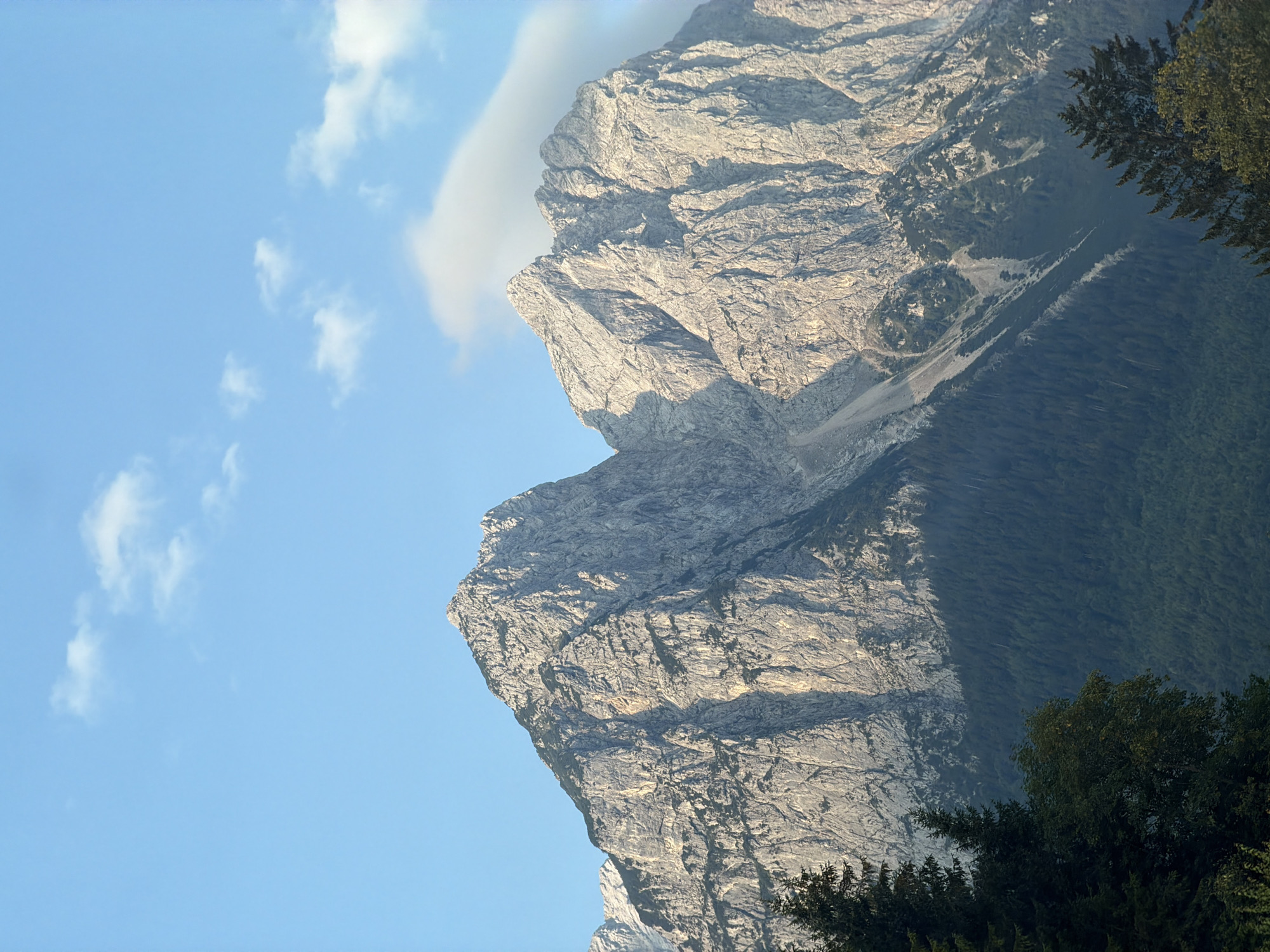 This is a vertical, medium shot of the jagged, sunlit peaks of the Alps, likely taken from the vicinity of Garmisch-Partenkirchen, Germany, given the characteristic mountainous landscape. The imposing, rocky mountains dominate the upper half of the frame, their surfaces textured with crevices and striations. Patches of dark green evergreen trees form a dense forest at the base of the mountains, transitioning to a lighter green and then the bare rock faces. A sliver of a third, further mountain range is visible on the far left, its summit partially obscured. The sky is a clear, bright blue with scattered, wispy white clouds. The lighting suggests it is daytime, with the sun casting a warm glow on the mountain peaks and creating shadows in their valleys. There are no people or animals visible in the image. The scene is a natural, outdoor environment characterized by its rugged terrain and vegetation.