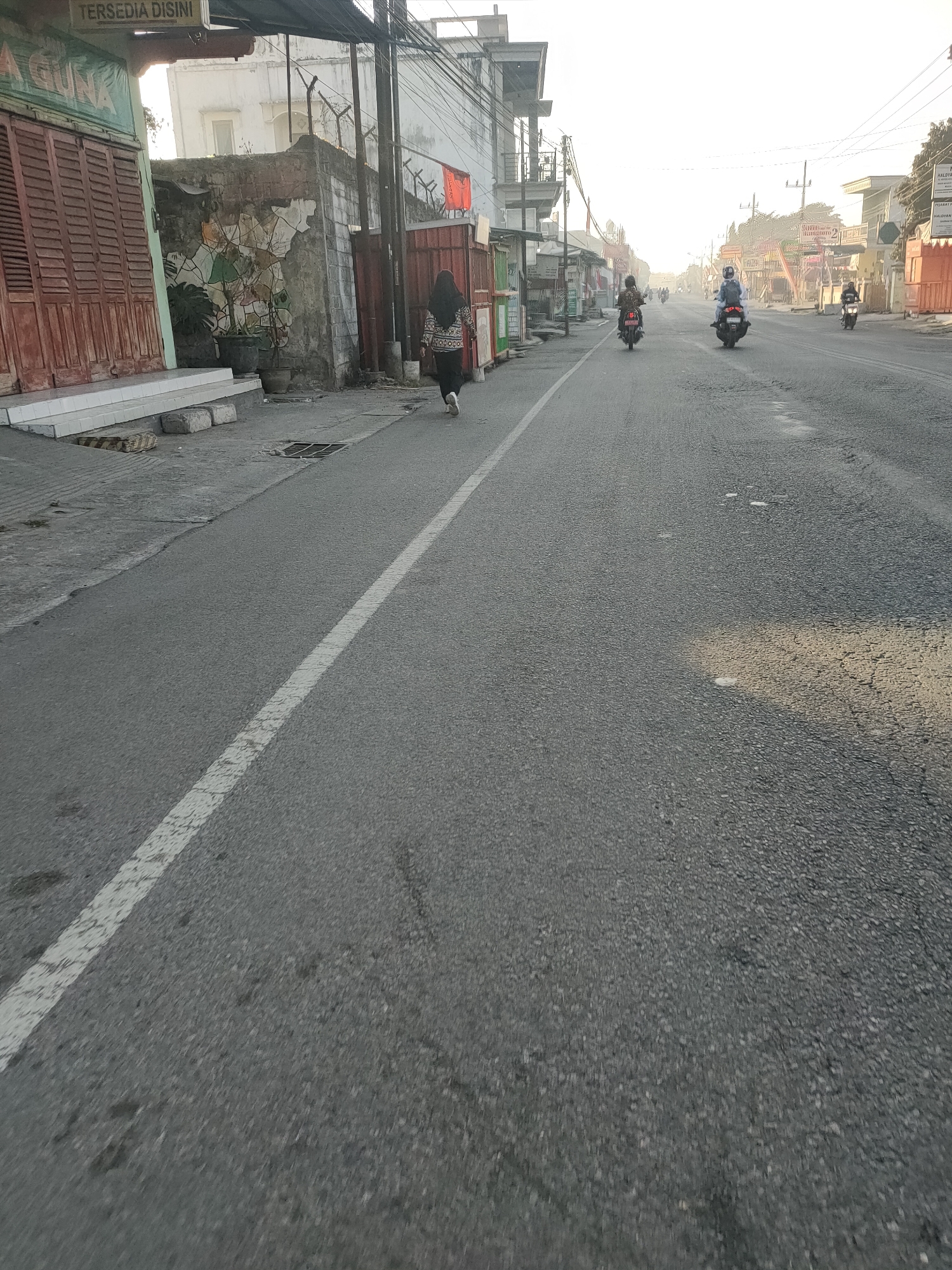 The image depicts a street scene in Kanigoro, Indonesia, likely during the morning. The road is paved and shows signs of wear, with visible potholes and a white lane marking. On the left side of the road, there are several buildings, including one with closed wooden shutters and another with a mural on its wall. A person wearing a hijab and a patterned top is walking away from the camera on the sidewalk. Further down the street, several motorcycles are visible, with riders wearing helmets. The background shows more buildings and trees, with a hazy, bright sky suggesting early morning light. The overall atmosphere appears to be that of a typical urban street in Indonesia, with everyday activity.