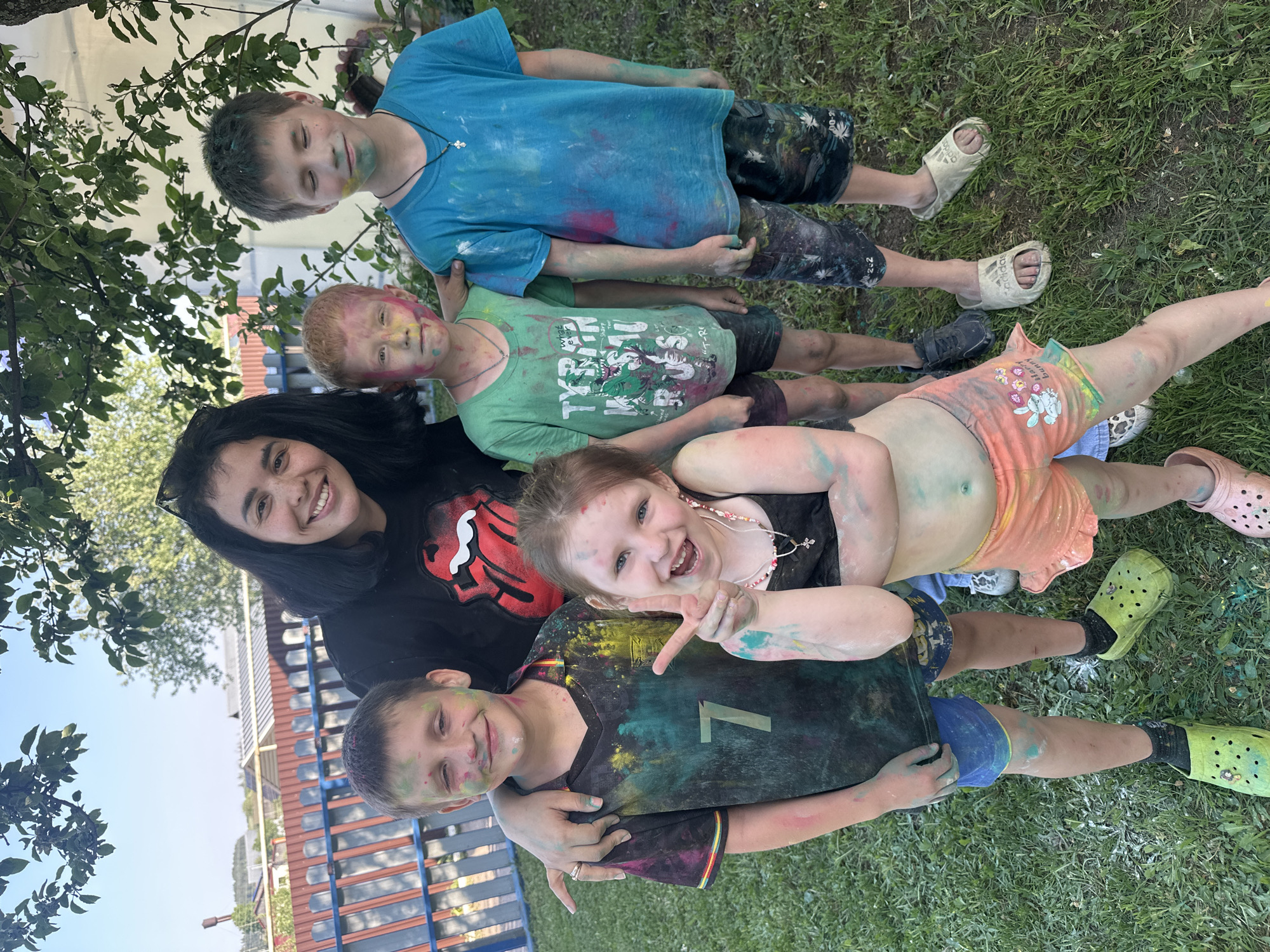 A group of five individuals, consisting of one young woman and four children, are lying on a grassy area outdoors. All individuals appear to be covered in colorful powder paint. The young woman is positioned on the left of the frame, smiling and wearing a black t-shirt with a Rolling Stones logo. The children are positioned to the right of her, forming a stack, all with visible smiles and some wearing necklaces. Each child is dressed in casual clothing, with varying colors and designs. The environment consists of green grass, with a building visible in the background. There is a tree with leaves partially obscuring the upper-left corner of the frame. Location is Unknown, Unknown.
