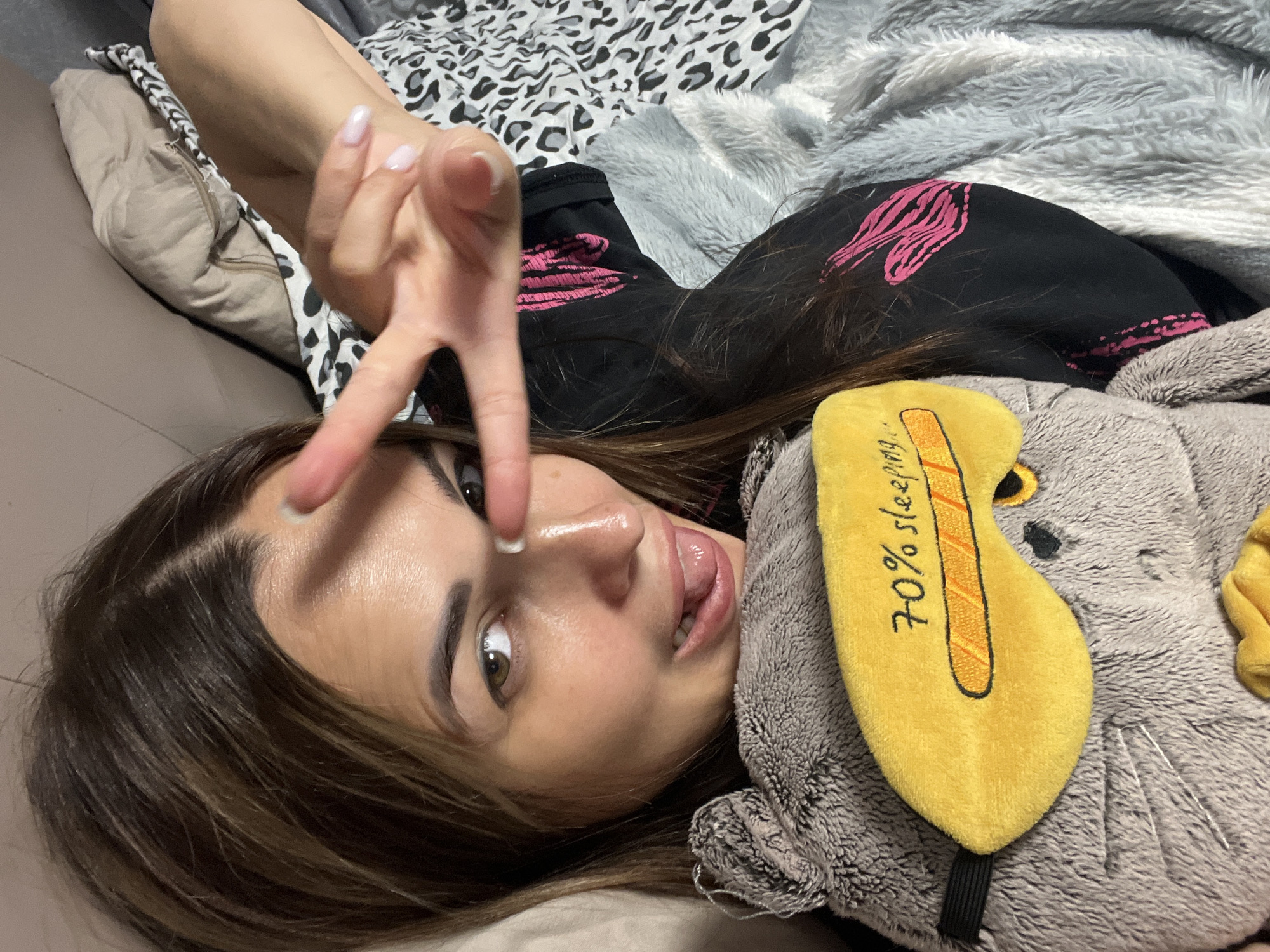 The media file features a close-up, overhead shot of a young woman lying down, possibly on a bed, as indicated by the presence of blankets and pillows. The woman is looking towards the camera with a playful expression, sticking her tongue out slightly. She has long, brown hair with lighter streaks and appears to be wearing black clothing with pink accents. Her left hand is raised, displaying a peace sign with painted nails.
Next to the woman is a stuffed animal, possibly an elephant, with a yellow eye mask resting on its head. The eye mask has a black band and is decorated with an orange stripe and black outline. Visible text on the eye mask reads "70% sleeping."
The overall scene suggests a casual and relaxed setting, likely indoors, given the soft lighting and bedding. The woman's pose and expression convey a sense of fun and informality. There are no clear indicators of the time of day, weather, or specific location beyond the domestic setting.
