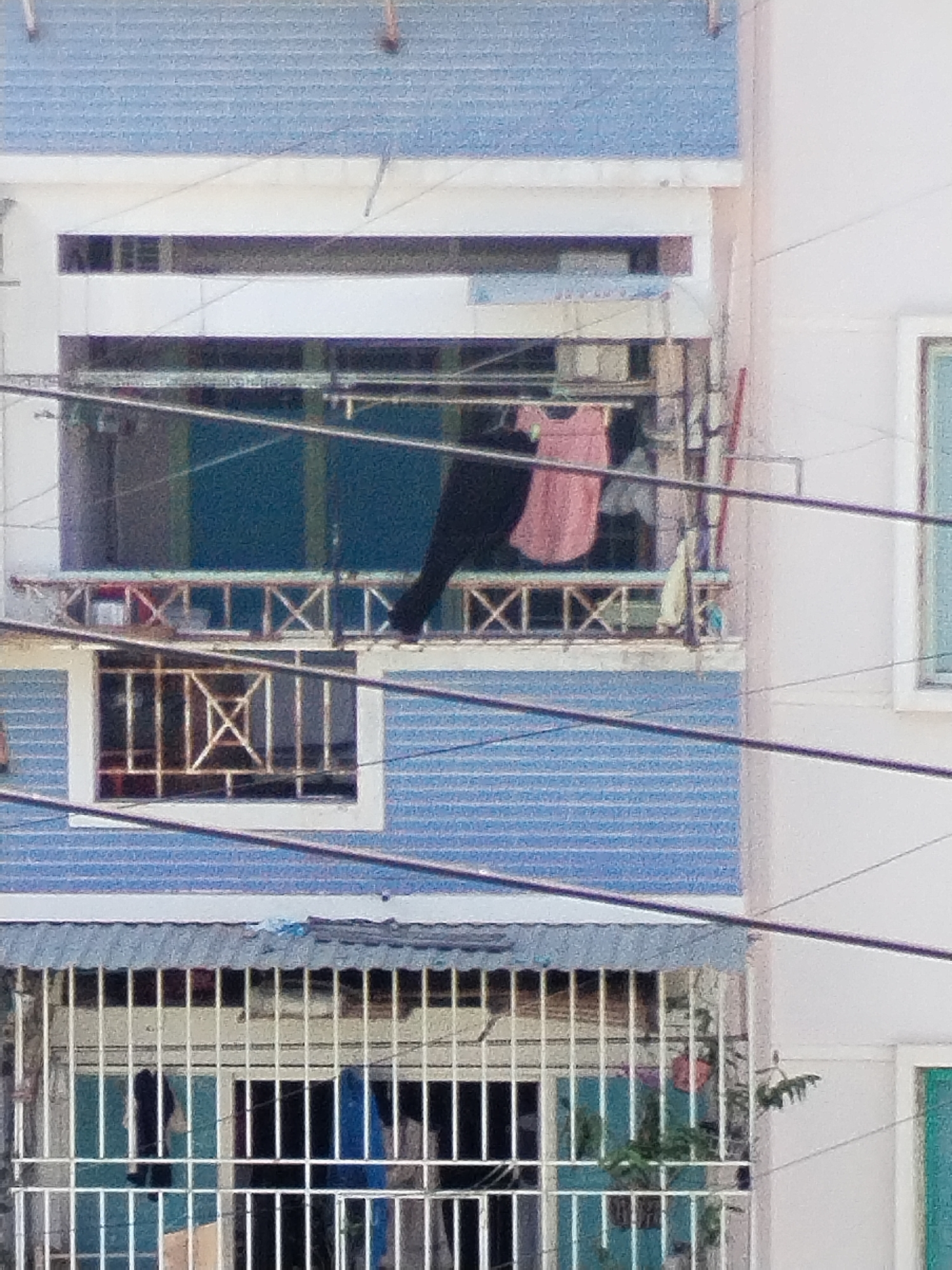 The image depicts a partial view of a building in Nha Trang Ward, Vietnam. The building features multiple levels with varying architectural designs. Laundry hangs drying on a balcony enclosed with a metal railing on the upper level. Below, another balcony is visible through vertical bars. Power lines stretch across the foreground, partially obstructing the view of the building. The building is painted in shades of blue and white. There are no people visible. Plant life is visible at the lower right, indicating the presence of vegetation nearby.