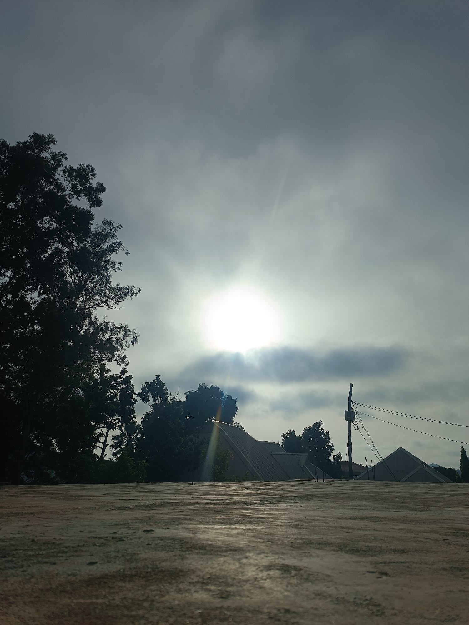 The image depicts an outdoor scene, likely taken from a rooftop in Bukuru, Nigeria. The focus is on the expansive cloudy sky with the sun obscured by the overcast conditions. The light from the sun creates a bright glare, with rays extending in multiple directions. In the midground, silhouettes of trees and buildings are visible. A utility pole with wires is also present on the right side. The foreground consists of a flat, concrete surface, which appears to be the rooftop from which the photograph was taken. The overall impression is that of a cloudy day in a semi-urban or rural setting. No people are visible in the frame.
