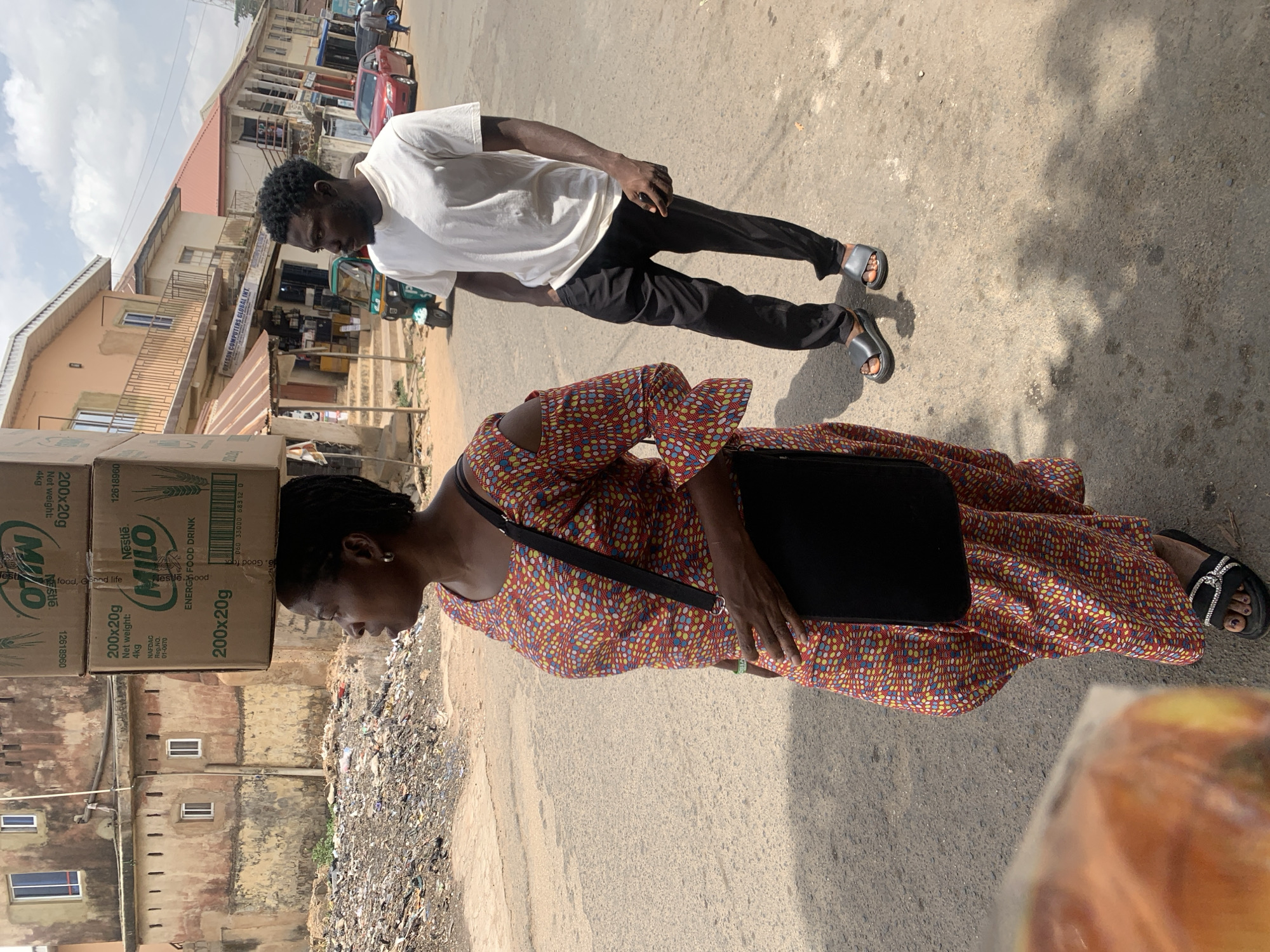 This image depicts a bustling street scene in Jos, Nigeria, under bright daylight with clear skies. In the foreground, a woman is seen walking while expertly balancing a stack of at least three large cardboard boxes on her head. The boxes are labeled "Nestlé MILO ENERGY FOOD DRINK," each indicating "200x20g" and "Net weight: 4kg," and feature "NAFDAC Reg.NO. 01-0670," confirming their Nigerian origin. She wears a vibrant, patterned dress in red, orange, blue, and yellow hues, black sandals with a shiny embellishment, and carries a black shoulder bag.
Behind her, a man walks in the same direction, dressed in a simple white t-shirt, dark trousers, and dark slide sandals. His dark hair is short, and his expression is neutral.
The setting shows a paved street with some dirt and debris, including a visible pile of trash along the side of the road near the woman. Buildings of varying heights and styles line the street, some appearing to be shops, and one prominently displays a sign for "OKESON COMPUTERS GLOBAL INT." A small green and yellow tricycle taxi (Keke Napep) and a red car are visible further down the road, indicating common modes of transport. The strong sunlight casts short, defined shadows, suggesting a midday or early afternoon time. Both individuals appear focused on their movement through the street.