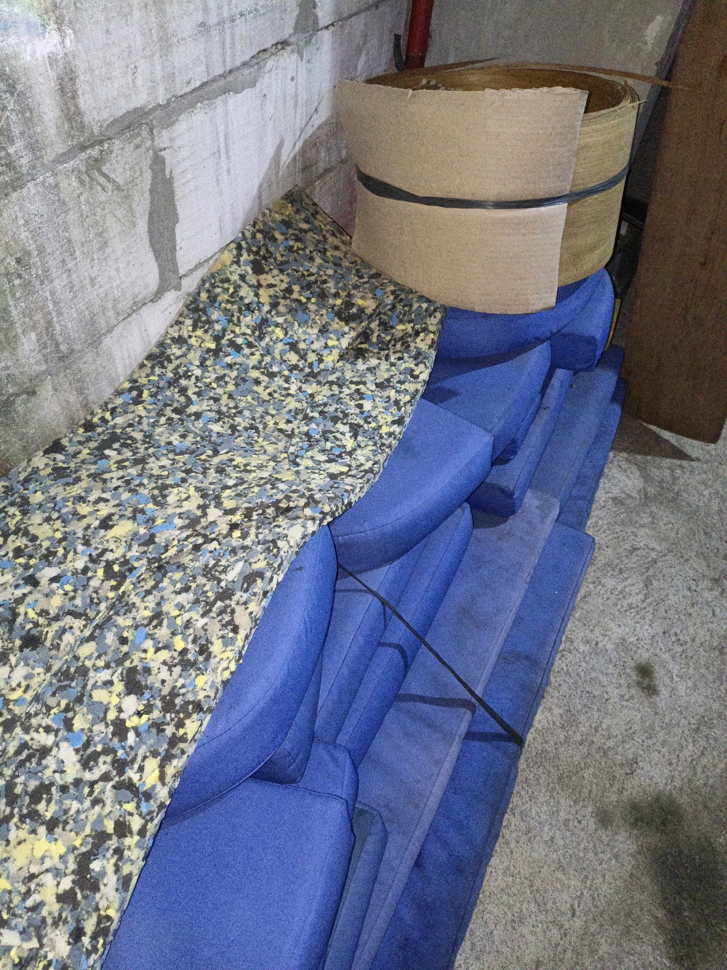 The image depicts a cluttered storage area. In the foreground, several pieces of blue foam padding are stacked. A large, rolled piece of composite board, secured with a black strap, rests on top of some of the blue padding. To the left, a piece of speckled foam material, likely from a cushion, is laid out. The background shows a concrete block wall and a concrete floor. The scene is lit, but shadows are present, suggesting artificial lighting. There are no people visible in the image. The overall impression is one of storage or disarray. The image does not provide any specific information about the location in the city of Cikal, Indonesia.