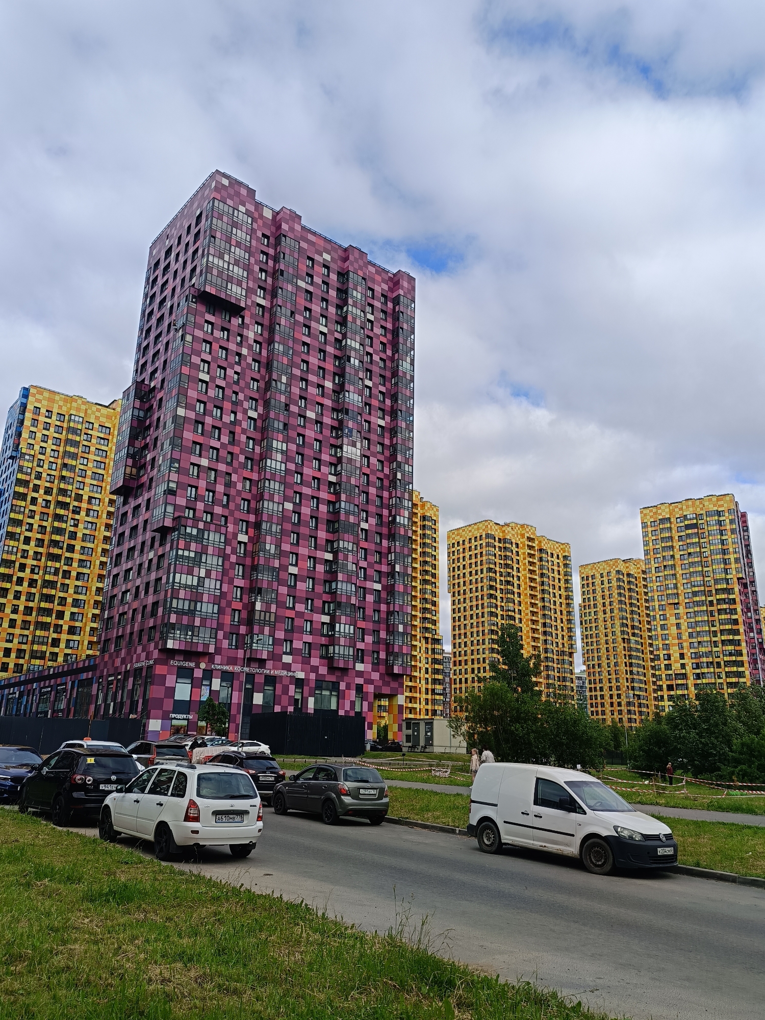 The image depicts a modern residential complex in Saint Petersburg, Russia, with a row of high-rise buildings, some colored in vibrant yellow and others in shades of pink and purple. The buildings feature a distinctive checkered facade pattern. In the foreground, a street lined with parked cars, including a white van, a white station wagon, and several dark-colored sedans, runs alongside a grassy verge. The sky is overcast with scattered clouds. A few trees are visible between the buildings and on the right side of the image. Signs on the pink and purple building are partially legible, indicating commercial establishments like a "Cosmetology and Medicine Clinic." There are a few individuals visible in the background near the buildings.