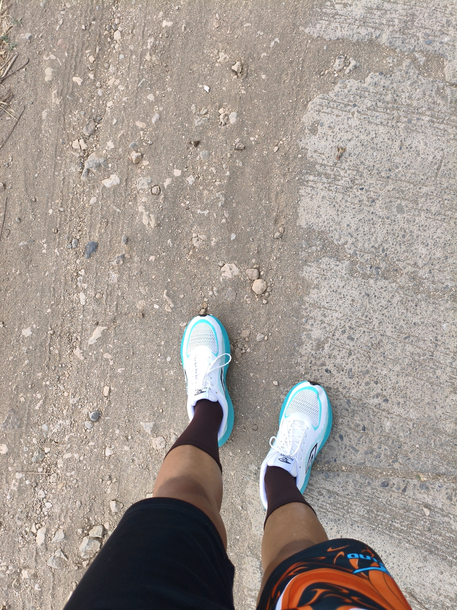 The image shows a person's legs and feet, wearing white running shoes with turquoise accents, dark brown socks, and black shorts. The ground consists of a mix of dirt, small rocks, and cracked concrete. The scene appears to be outdoors, possibly on a path or road. The environment suggests a rural or less urbanized area, potentially a trail or an unpaved road. The lighting indicates it is daytime.