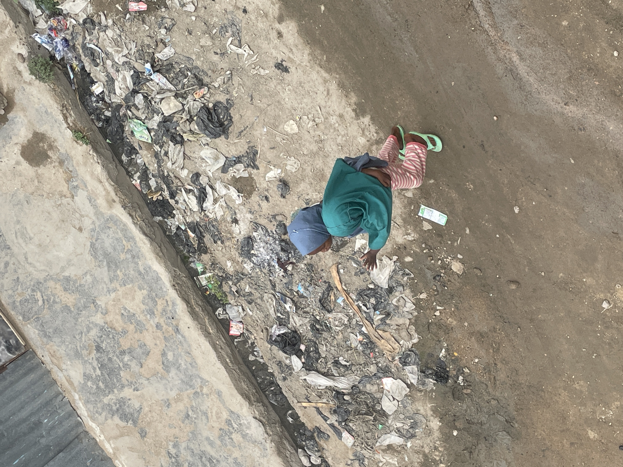 A person wearing a blue head covering, green top, striped pants, and green sandals is picking through trash in a dirty, trash-filled ditch. The person is located in an unspecified area of Unknown, Unknown. The ditch is full of various types of trash, including plastic, paper, and black bags. The background is composed of dirt, mud, and concrete.