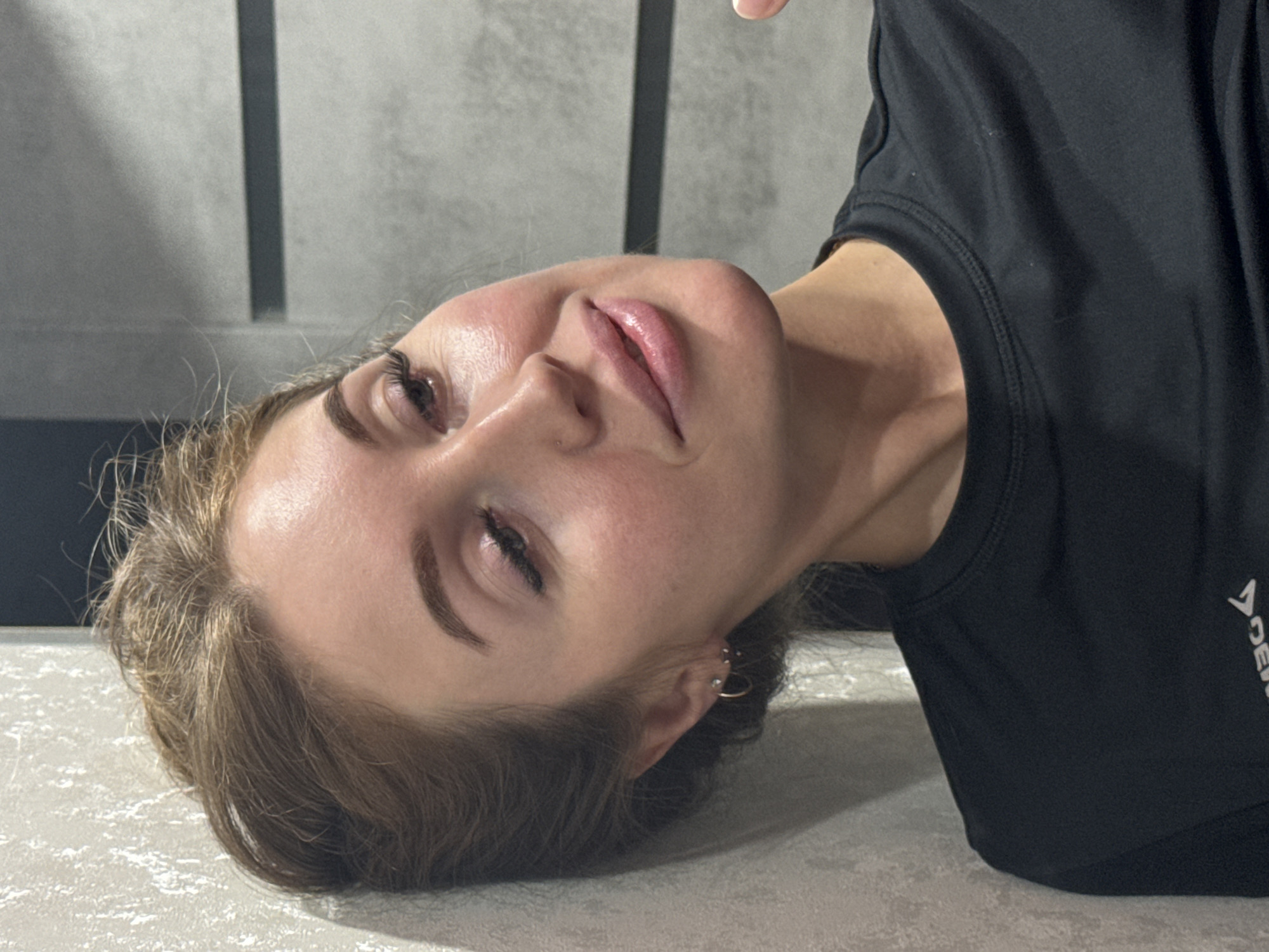A medium close-up portrait shows a young woman lying down or leaning back, her head tilted significantly to her left. She looks upwards towards the camera with a relaxed, slightly smiling expression, her eyes partially closed. Her light brown hair is spread around her head on a light, textured surface.
She has well-groomed eyebrows, subtle eye makeup, and pink, slightly parted lips. Her visible ear displays multiple piercings, including a small hoop and several studs or barbells. She is dressed in a black short-sleeved t-shirt, with a partial white angular logo or symbol visible on the right side. The background features a grey wall with distinct vertical panels. The scene appears to be indoors in a casual setting. The woman conveys a relaxed and content demeanor. The location context for this image is Chaikovsky, Russia.