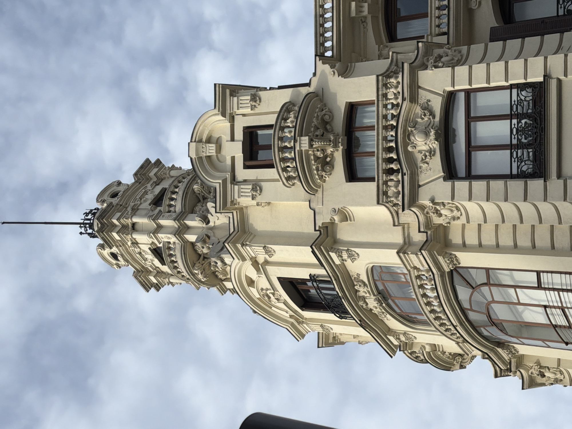 The image depicts a detailed, low-angle view of a building facade in Madrid, Spain, showcasing ornate architectural elements such as decorative moldings, elaborate window frames, and carved details. The building's facade is off-white or light beige, emphasizing the intricate baroque and neoclassical designs. Windows of various sizes and shapes are visible, some with decorative iron grilles. The sky is overcast, appearing light gray, with patches of clouds. The perspective emphasizes the verticality of the building, which extends upwards out of frame.