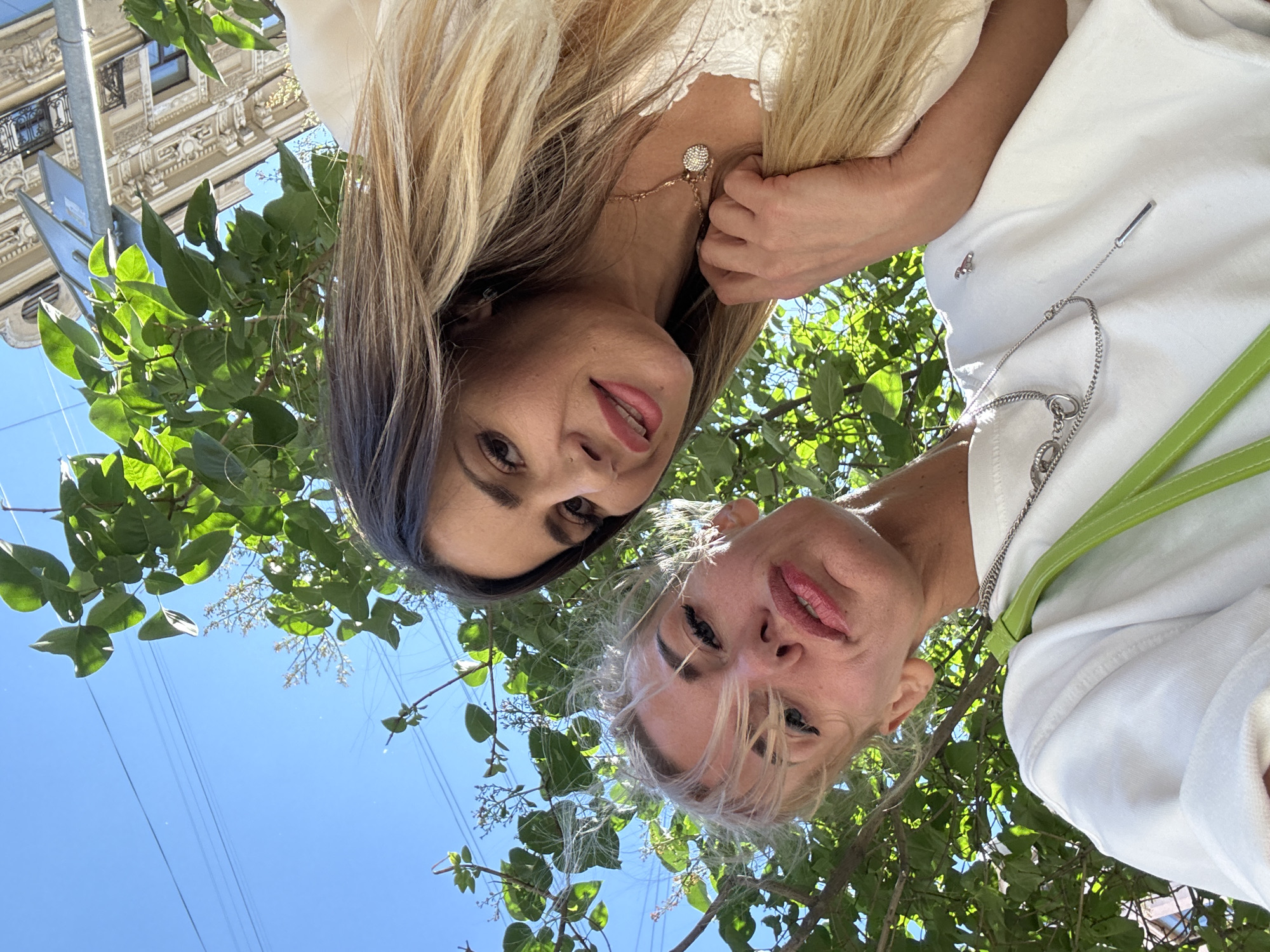 The media file is a close-up, low-angle selfie taken outdoors, featuring two women among tree branches with a building in the background. The woman in the foreground has blonde hair and is wearing a white top with a green strap over her shoulder and a silver necklace. She is smiling, with her lips slightly parted, and her eyes are looking directly at the camera. The woman behind her has long, dark blonde hair, and is wearing a white garment with lace detailing and a delicate necklace. She is also looking at the camera with a slight smile.
The scene is set on a clear, bright day, indicated by the blue sky and the strong sunlight casting shadows on the building and leaves. The building visible in the background appears to be of classical architecture, with ornate details and balconies, suggestive of an urban environment. The presence of tree branches in the foreground indicates a location with greenery, possibly a park or a street lined with trees. The orientation of the selfie suggests the subjects are looking upwards, perhaps posing for the camera. The overall mood is cheerful and casual, indicative of friends enjoying a sunny day. No visible text is present in the image. The context of Saint Petersburg, Russia, implies this scene is within that city.